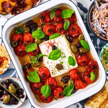Baked Feta with tomatoes in a white baking dish. Olives, hummus, slaw and bread surround it.