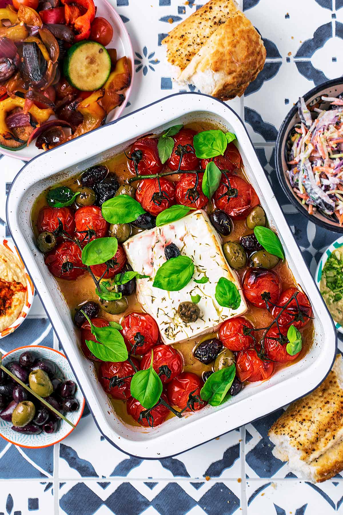 A block of cooked feta in a baking dish with cherry tomatoes, olives and basil leaves.
