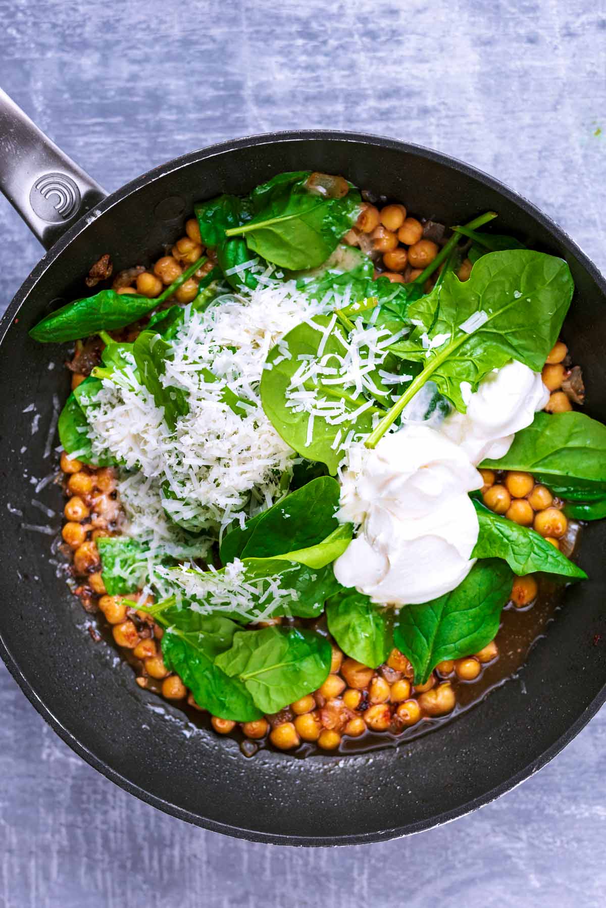A frying pan containing onions, chickpeas, spinach, cream and cheese.
