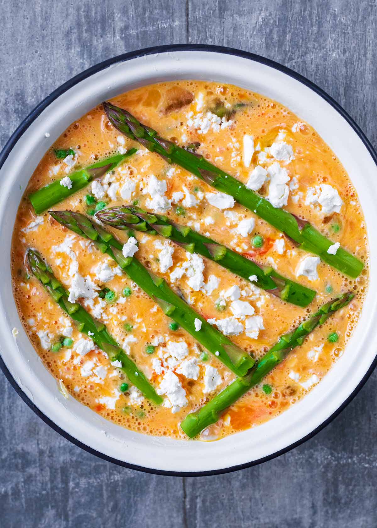 A baking dish containing whisked egg, asparagus spears, peas and crumbled feta.