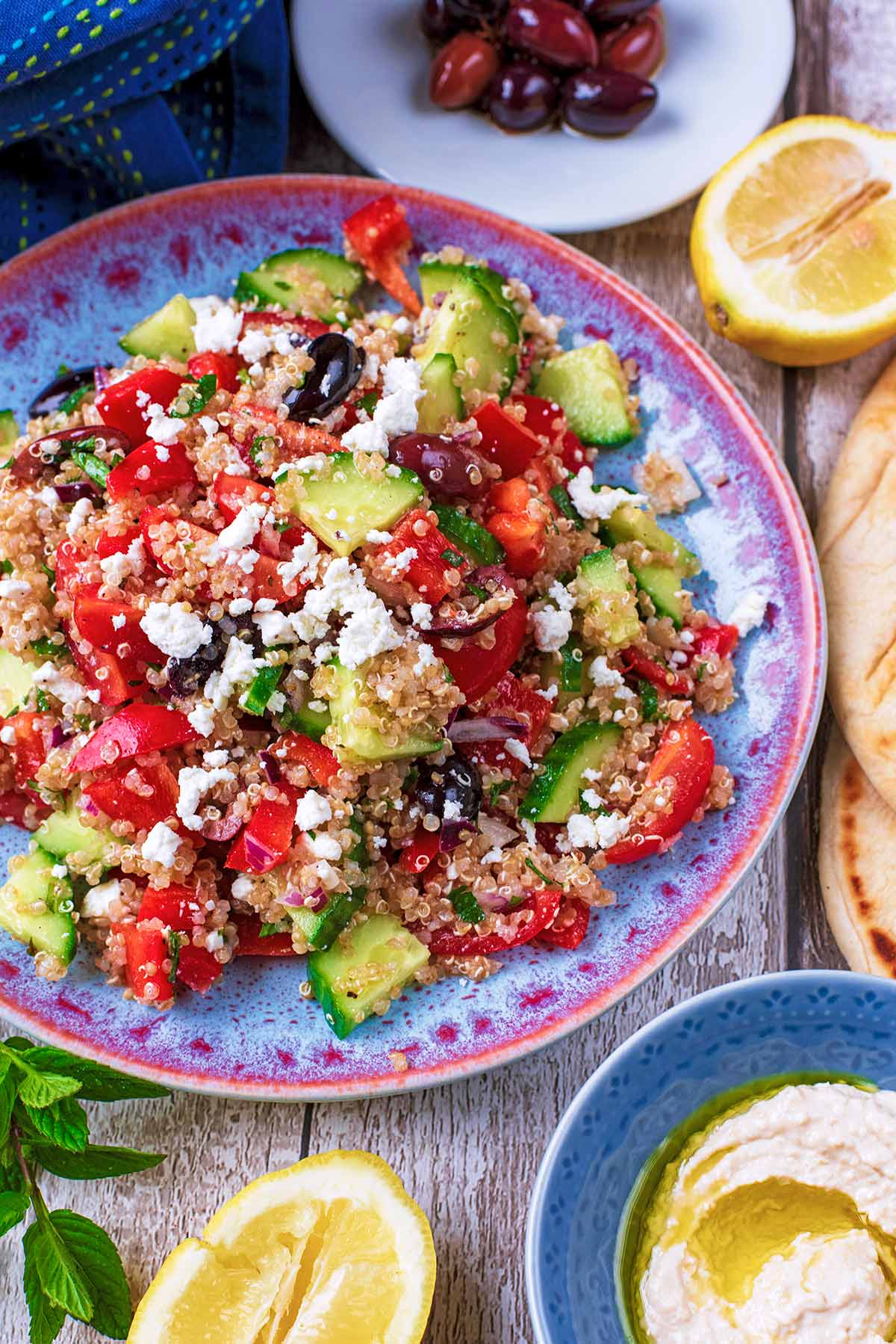 A plate of Quinoa Greek Salad next to small dishes of olives and hummus.