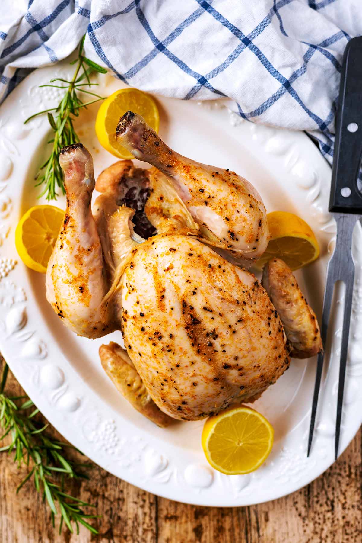 A whole cooked chicken on a large platter with lemon halves and rosemary sprigs