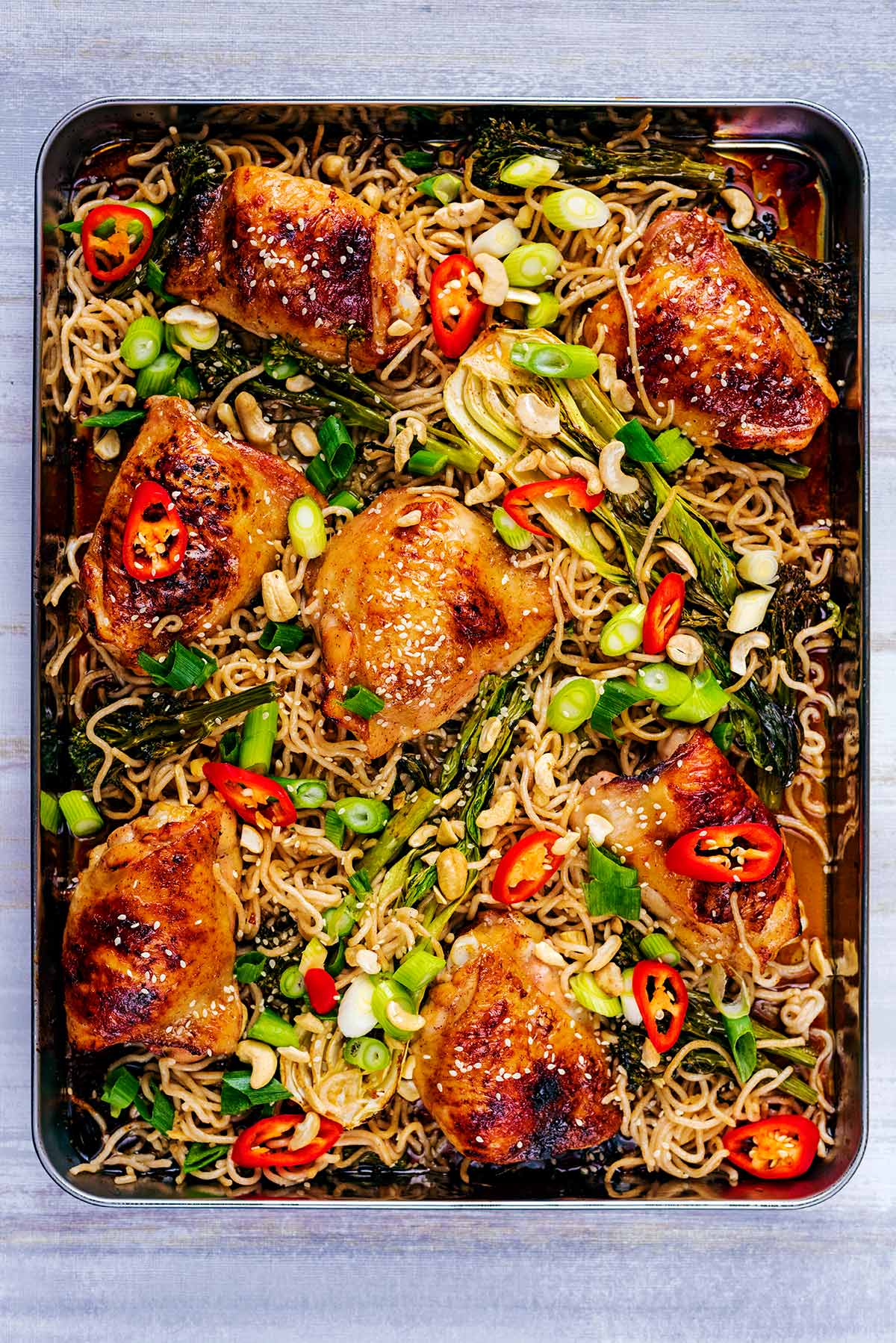 cooked chicken thighs and bok choy on a large baking tray with scallions, sliced red chillies and sesame seeds.