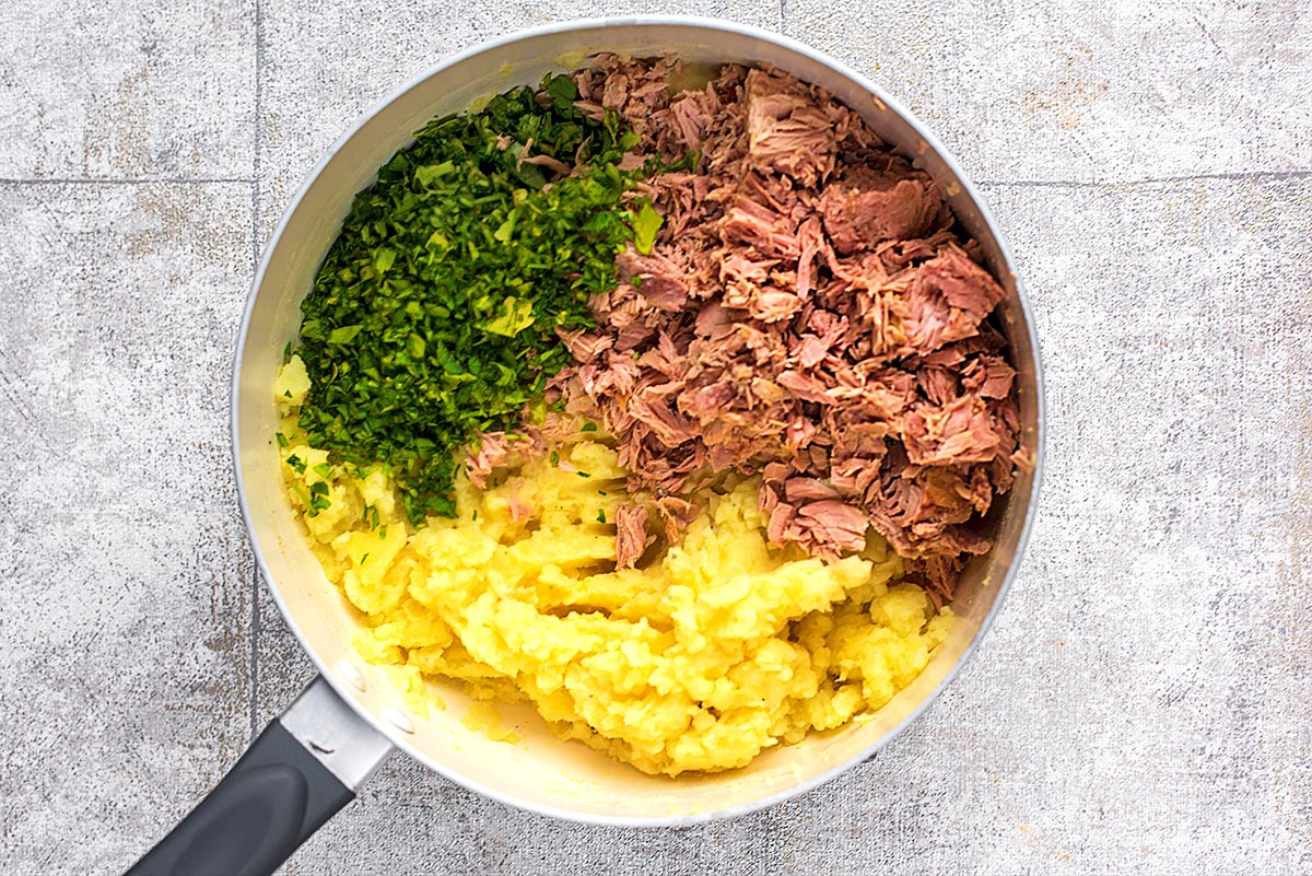 A saucepan with mashed potato, flaked tuna fish and chopped herbs.