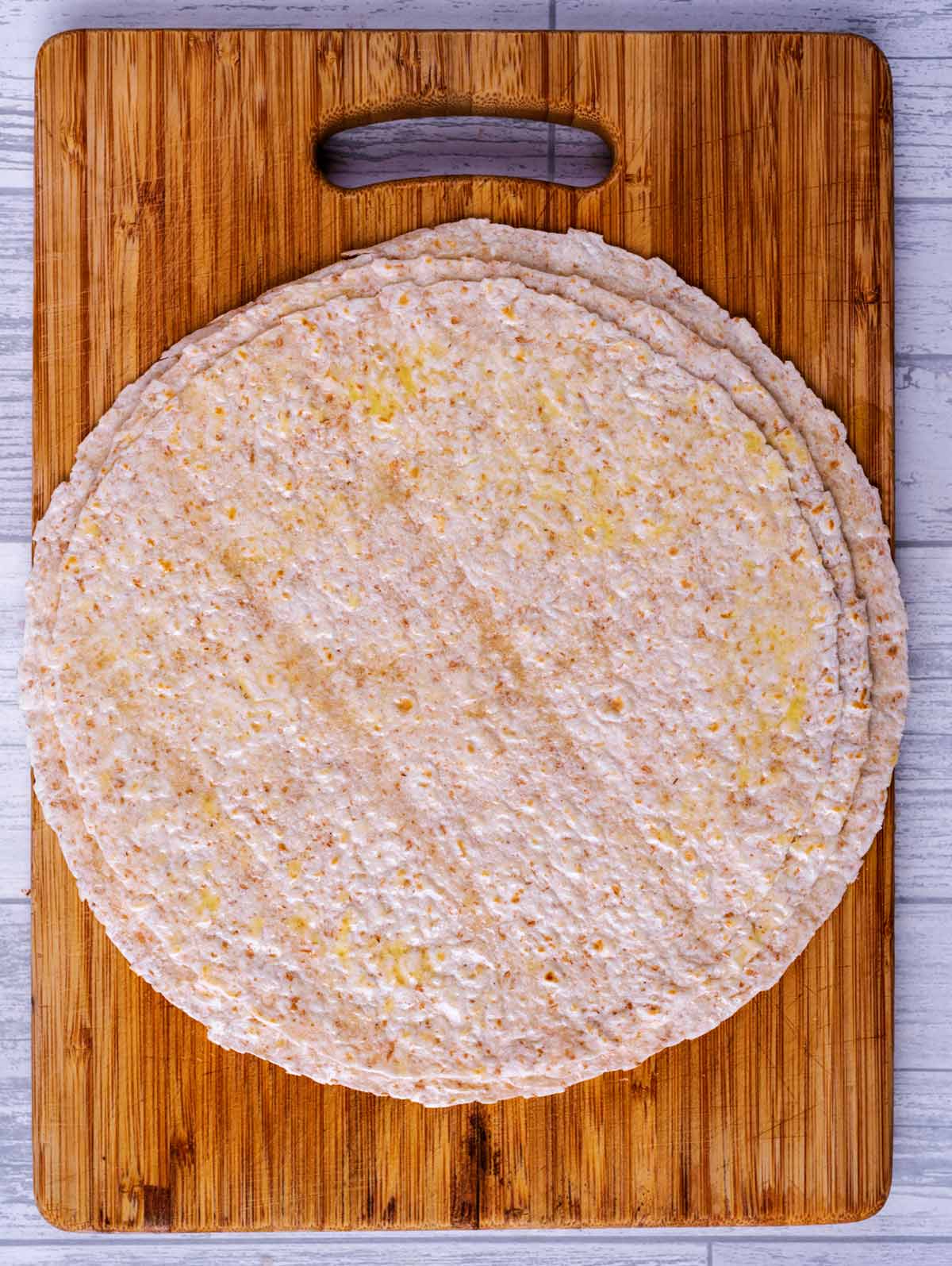 Flour tortillas spread with oil on a wooden chopping board.