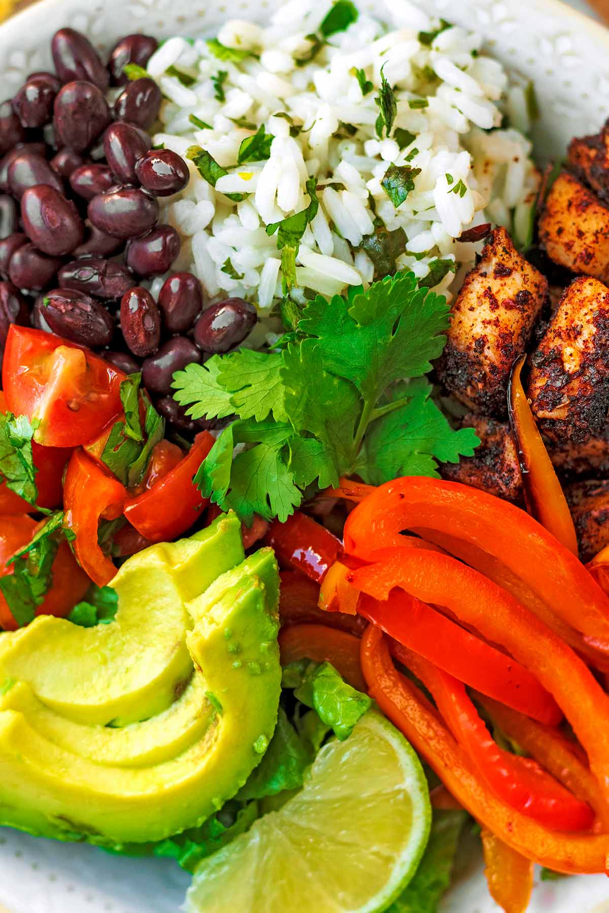 Sliced avocado, chopped tomatoes, black beans, cooked rice, seasoned chicken chunks, sliced red pepper and a lime wedge, all in a bowl together.