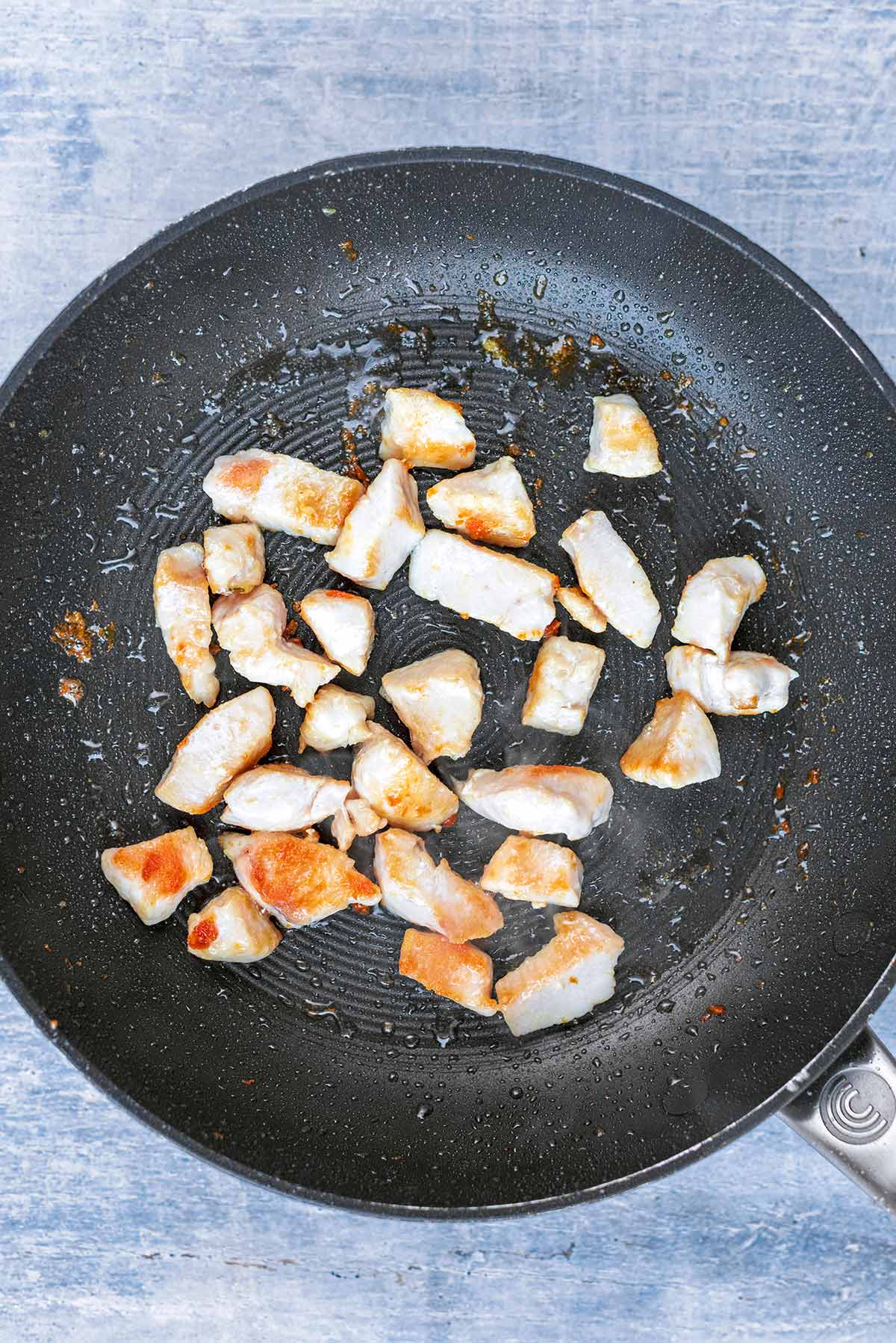 A frying pan with chunks of chicken cooking in it.