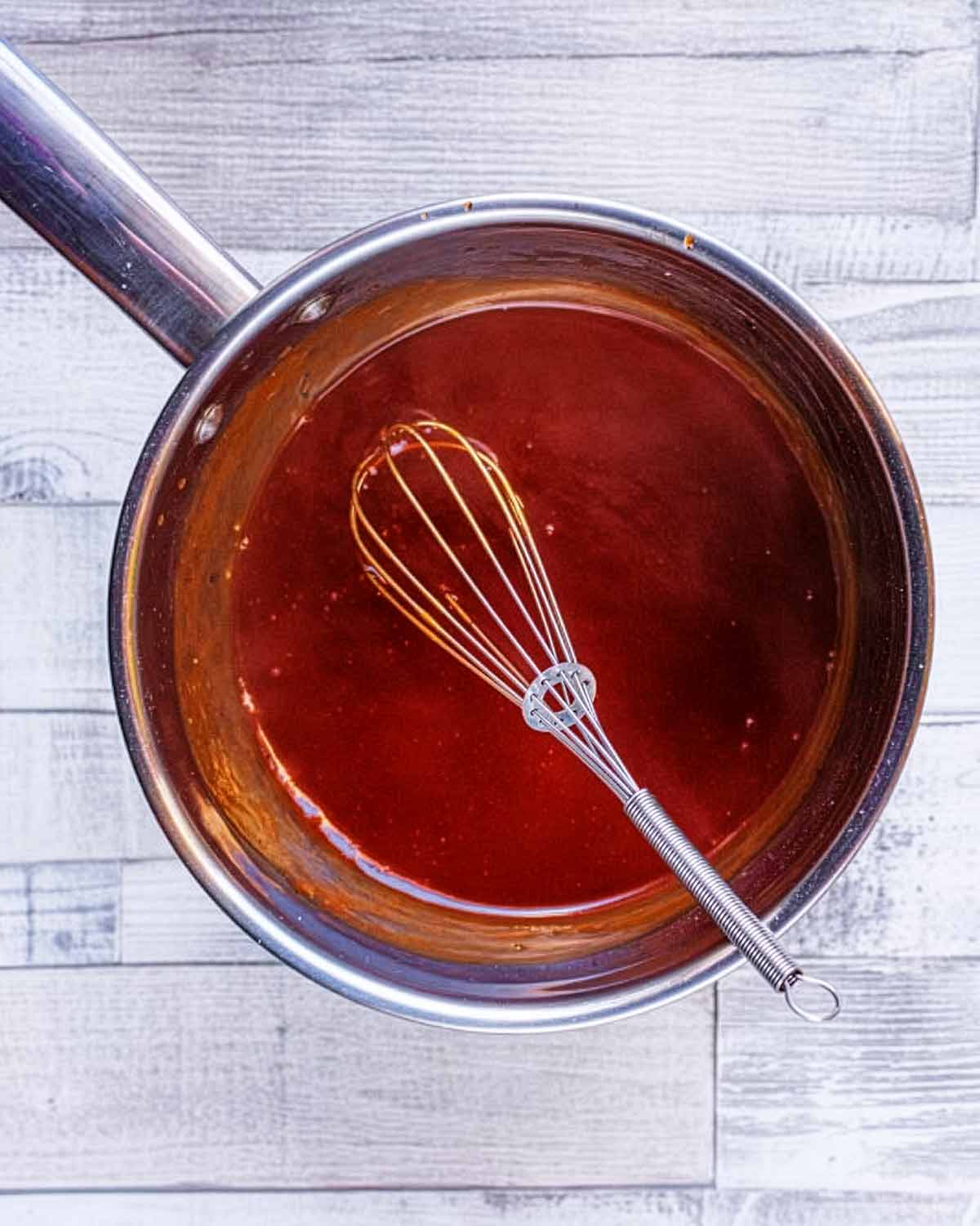 A saucepan containing sweet and sour sauce and a small whisk