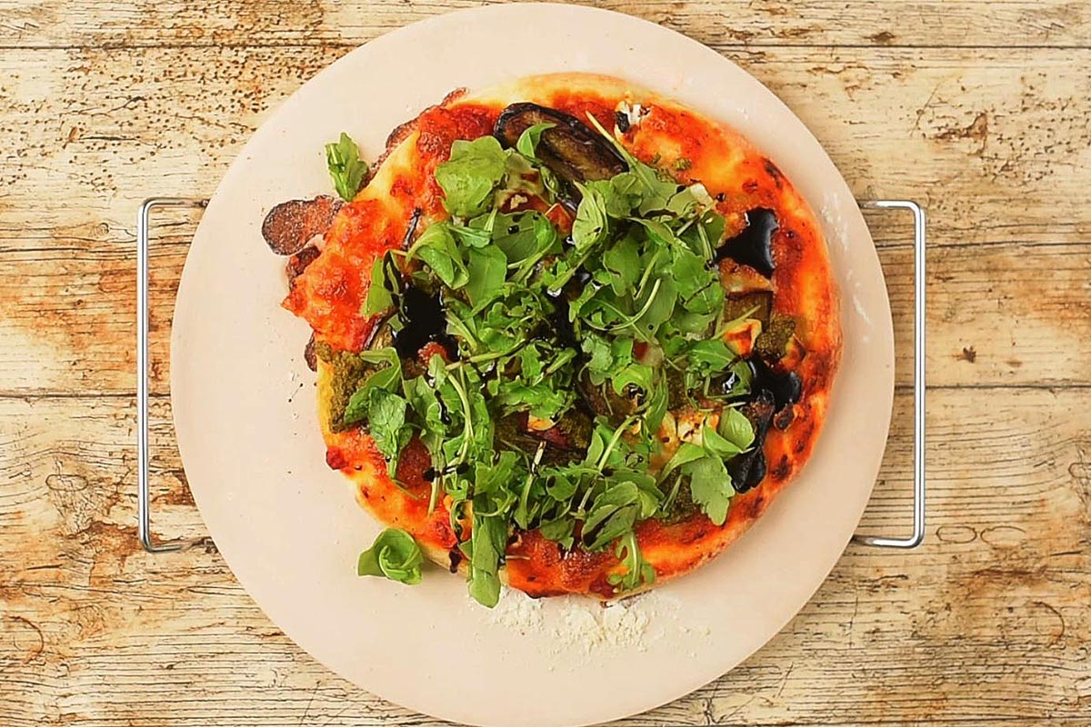 A cooked aubergine pizza topped with arugula and balsamic glaze.