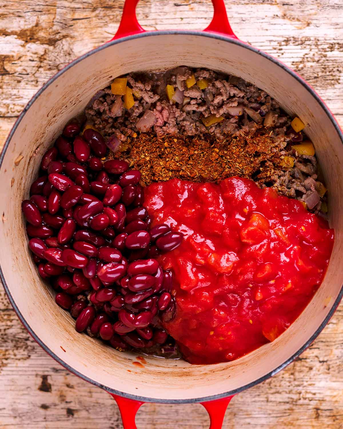 Cooked beef mince, red kidney beans, chopped tomatoes and spices in a large red pot.