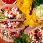 Salmon tacos arranged on a wooden board surrounded by tortilla chips and bunches of herbs.