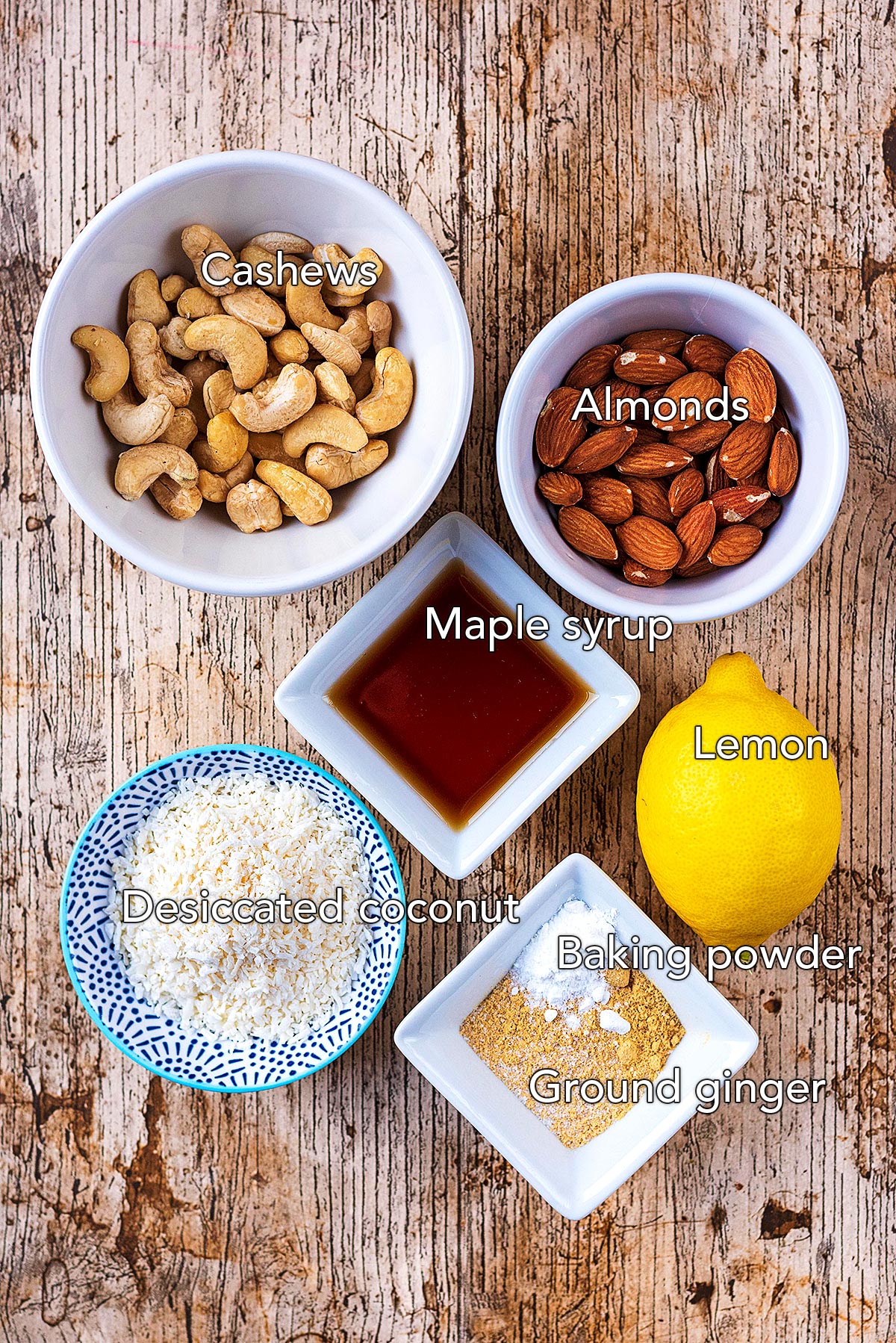 Small bowls of cashews and almonds, dishes of maple syrup, desiccated coconut and ground ginger and a lemon, all on a wooden surface.