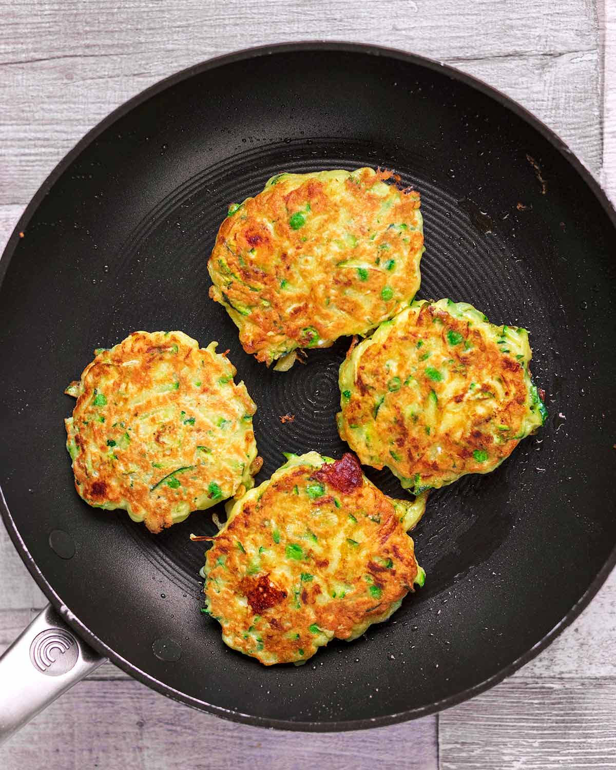 A frying pan with four courgette fritters cooking in it.