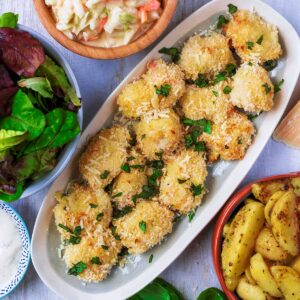 A plate of parmesan chicken bites next to a bowl of salad, some coleslaw and potato wedges.