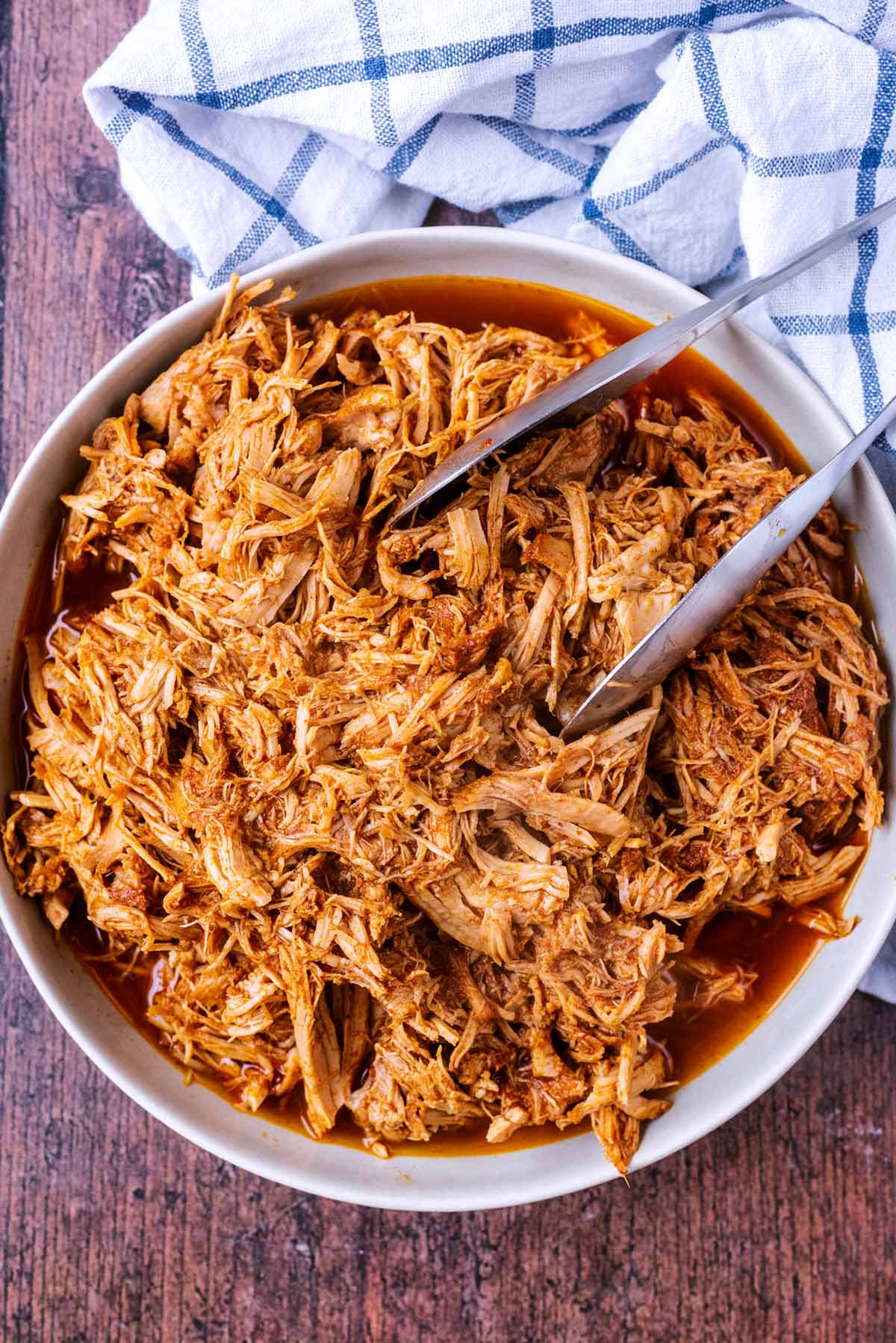 A large bowl of pulled pork with a pair of tongs in it.