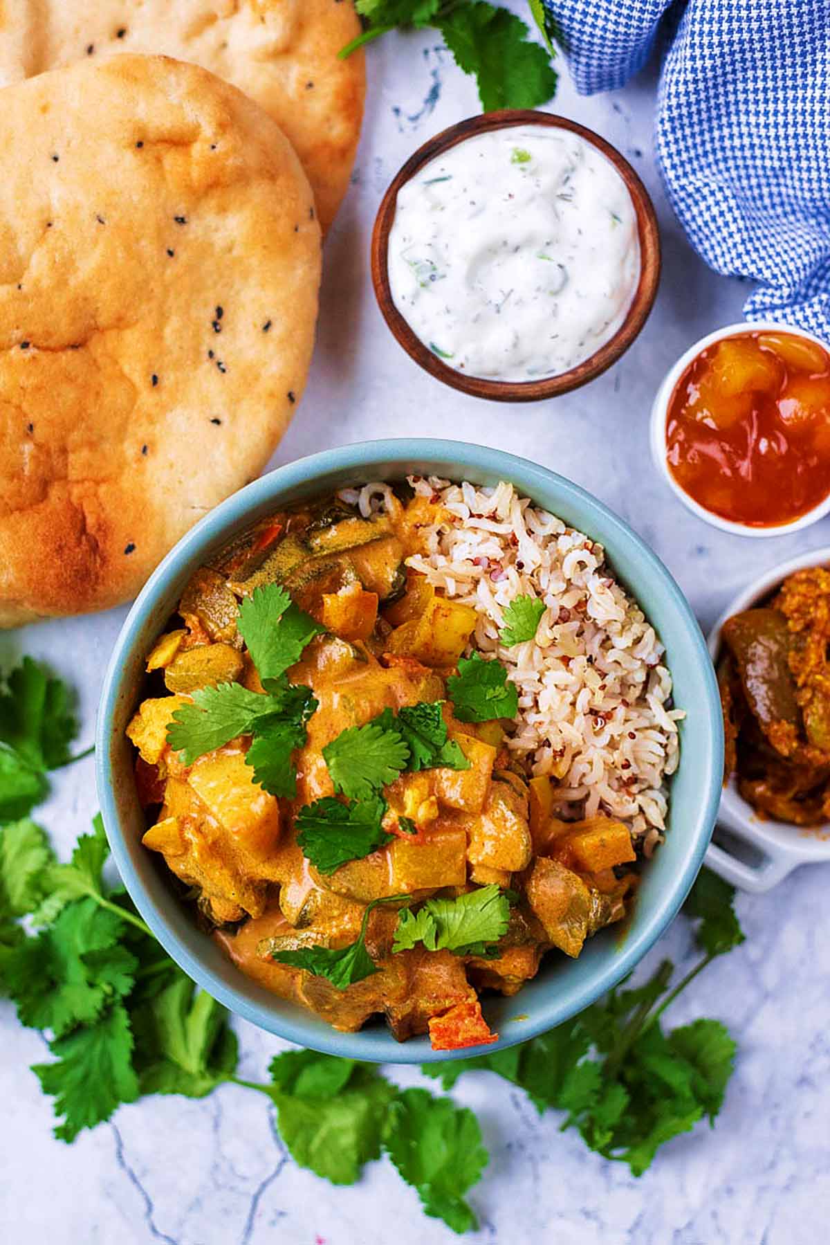 Vegetable curry in a blue bowl. Naan breads, raita, pickles and chutney sit next to it.