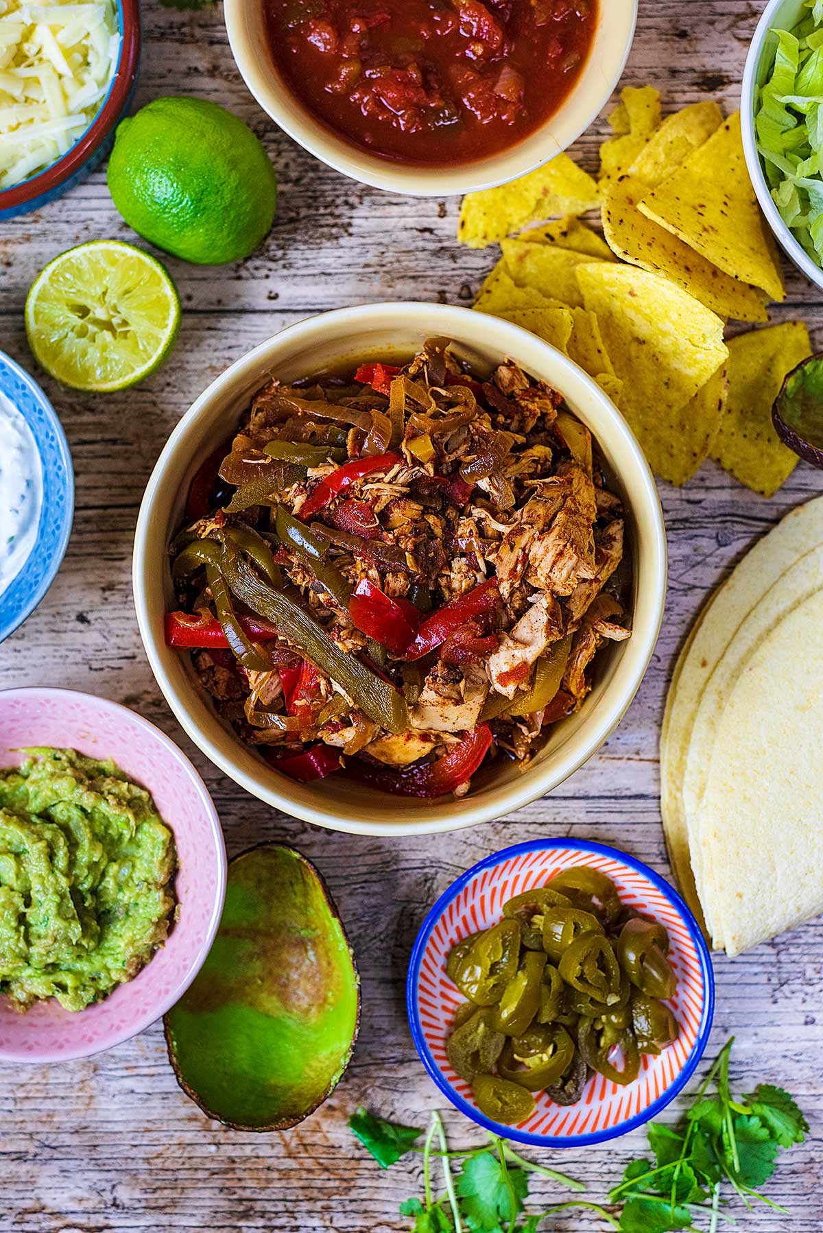 A bowl of fajita filling surrounded by salsa, guacamole, tortilla wraps, jalapenos and tortilla chips