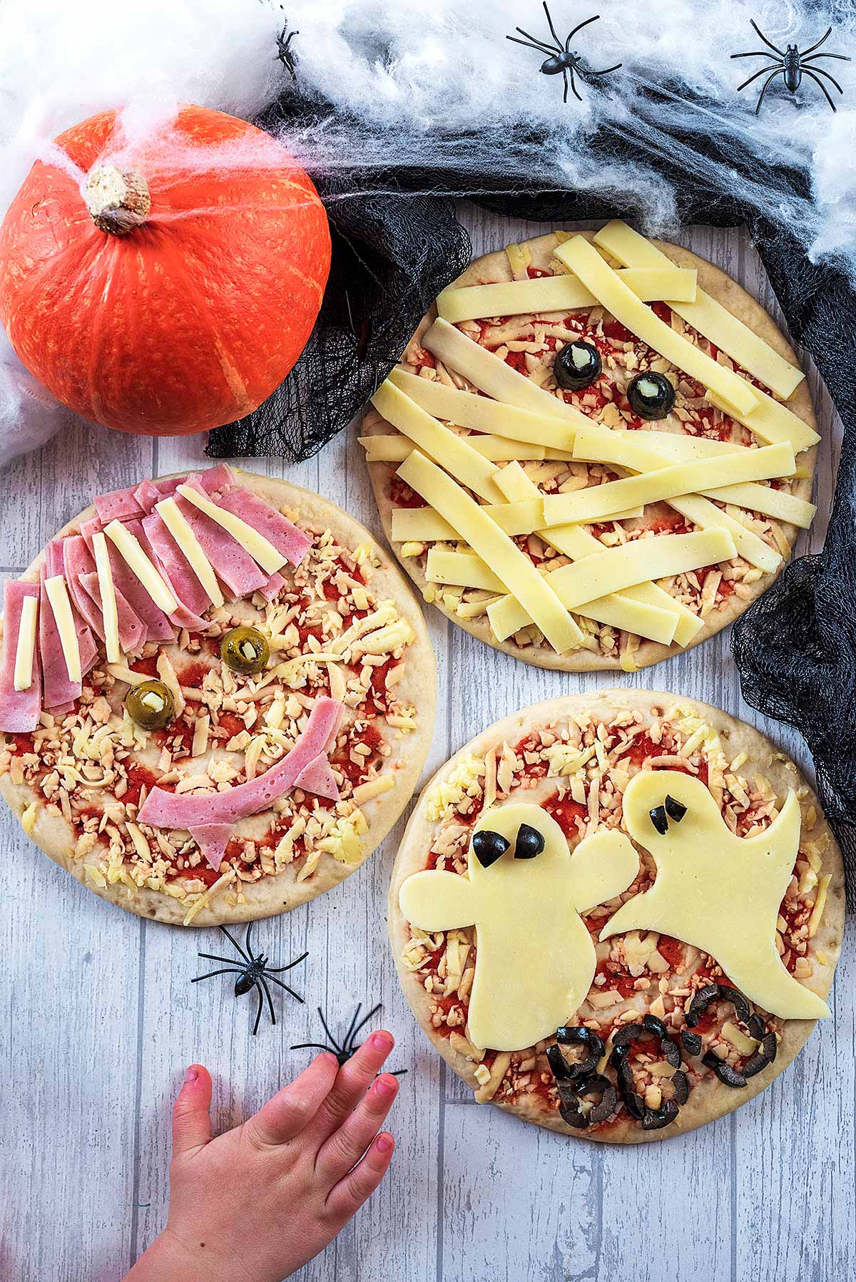 Three pizzas with Halloween themes. A child's hand is reaching in.