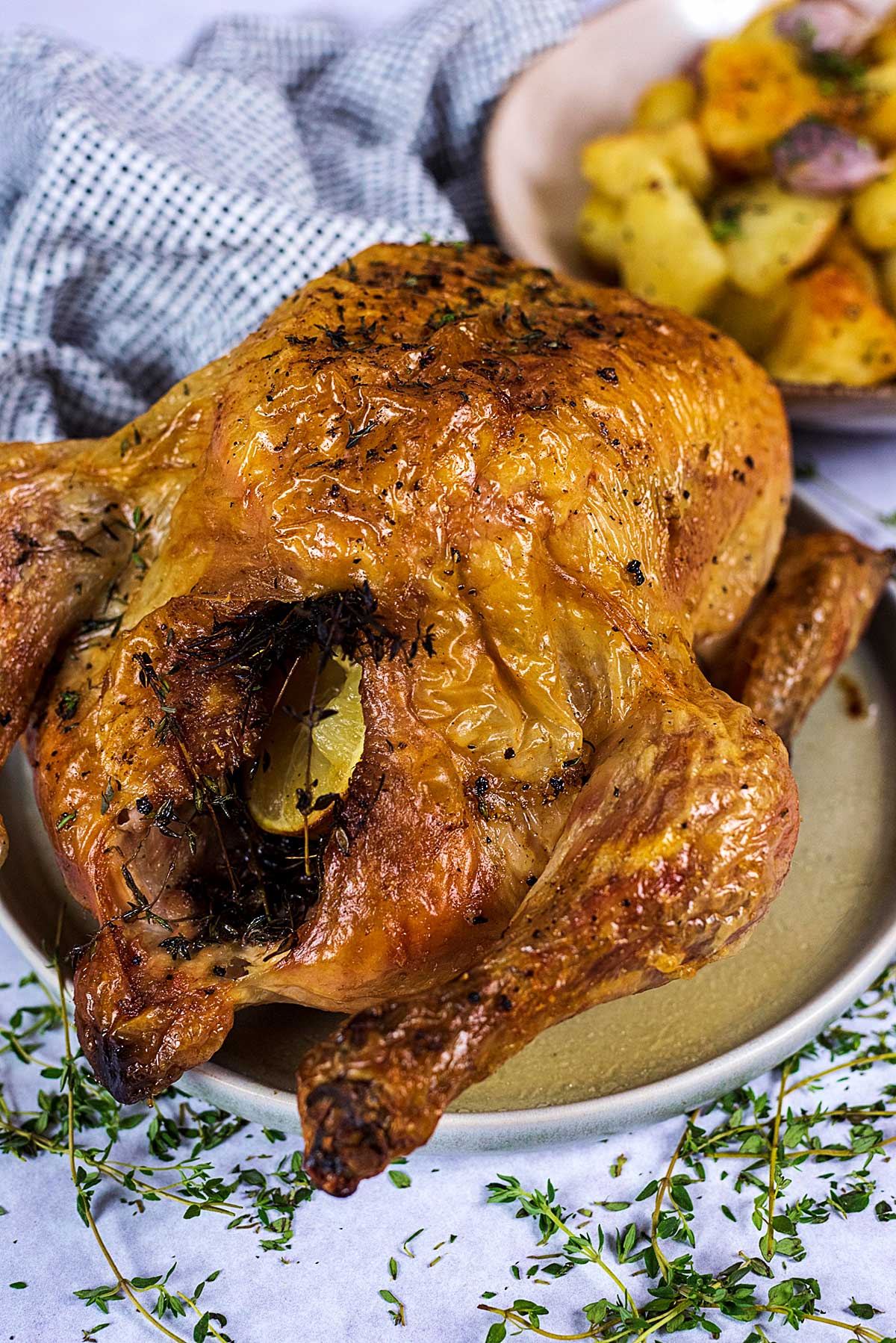 A whole roast chicken on a plate in front of a bowl of roasted potatoes.