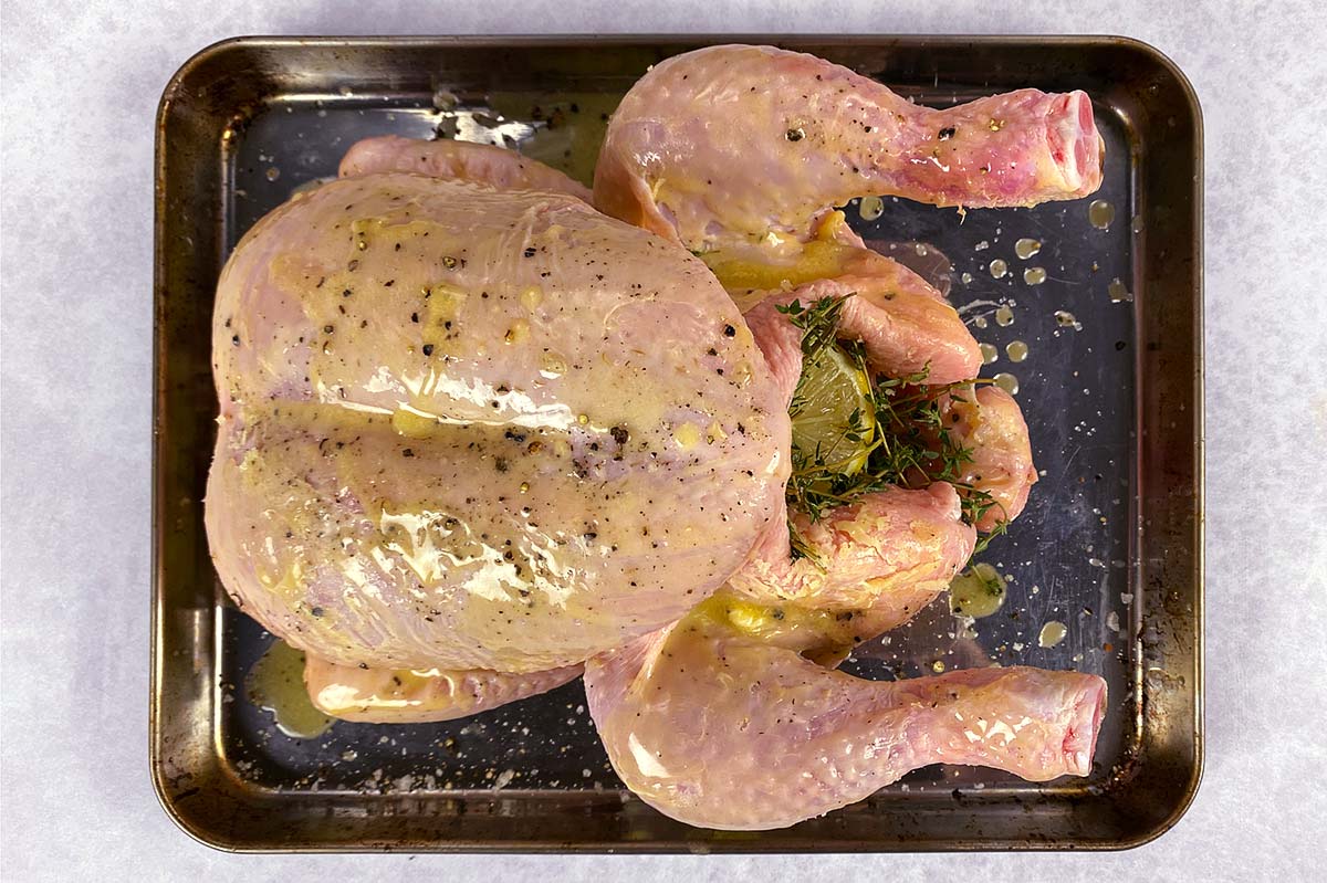 A whole uncooked chicken on a baking tray with lemons and thyme in its cavity.