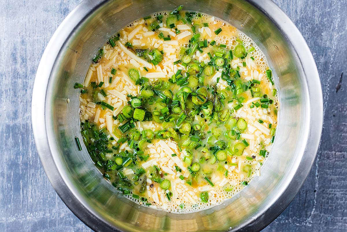 A metal mixing bowl containing whisked eggs, grated cheese, sliced spring onions and chopped chives.