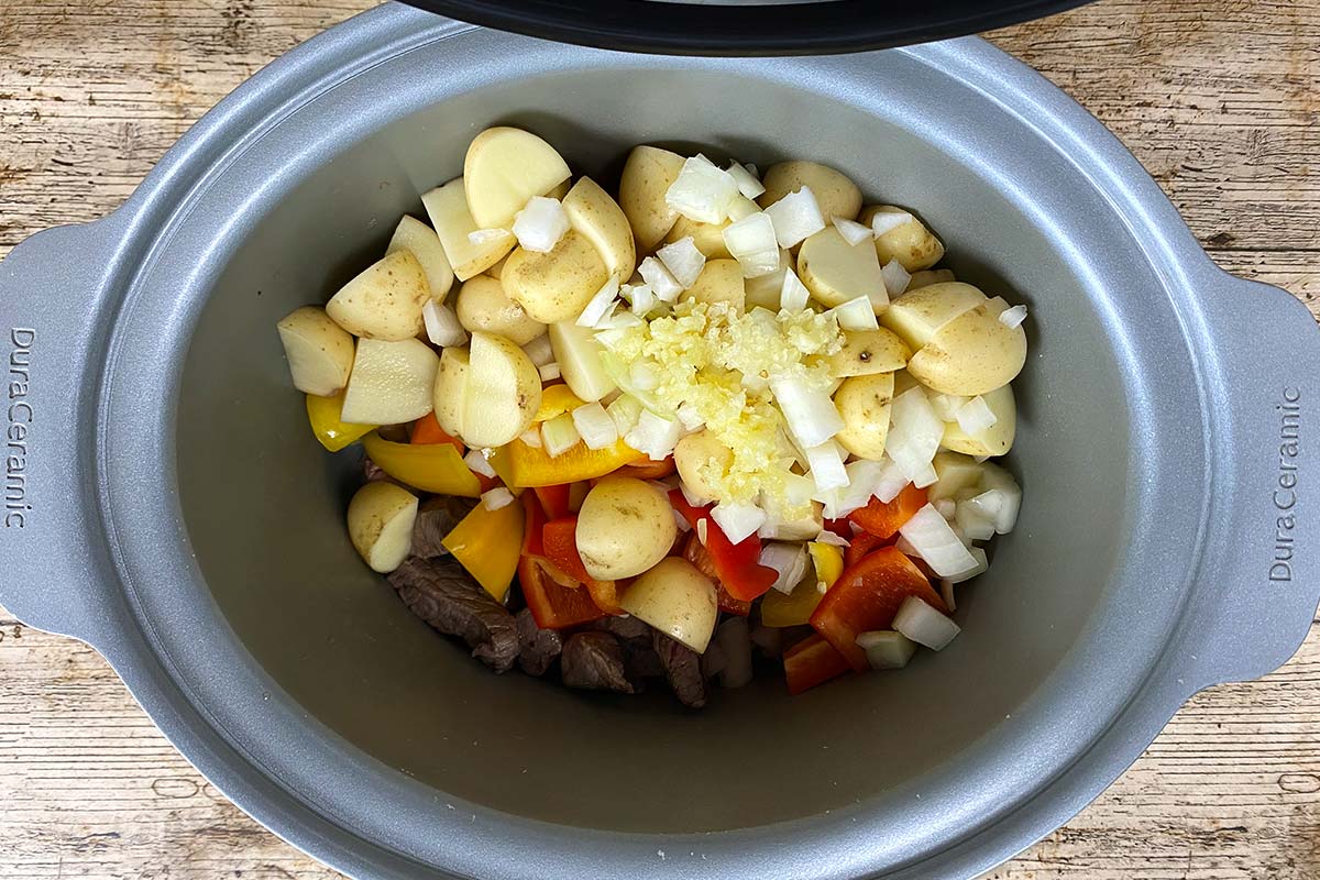 A slow cooker bowl containing browned beef and chopped onions, peppers and potatoes.