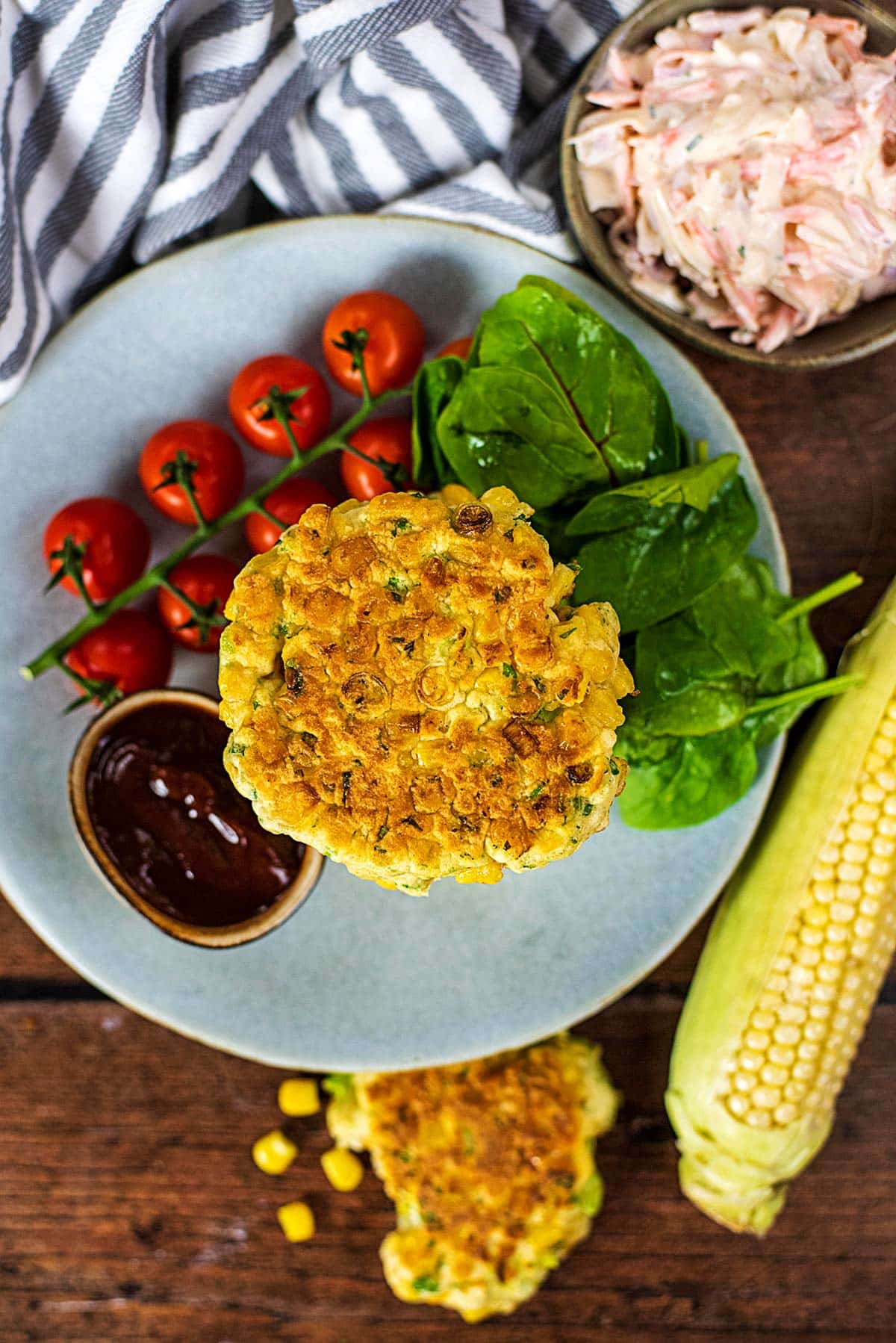 Sweetcorn fritters on a plate with cherry tomatoes, salad leaves and a pot of dip.