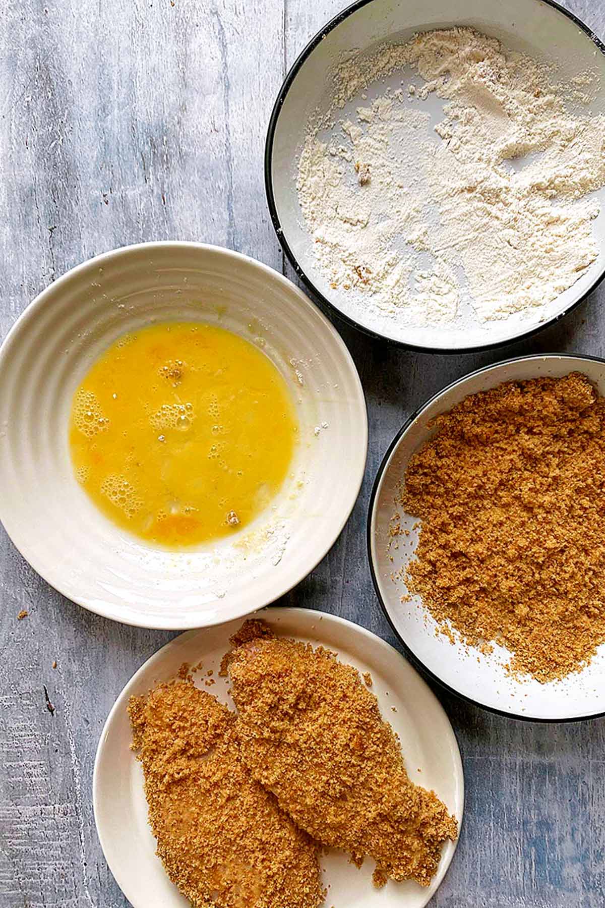 Bowls of flour, whisked egg and breadcrumbs next to coated chicken breasts.