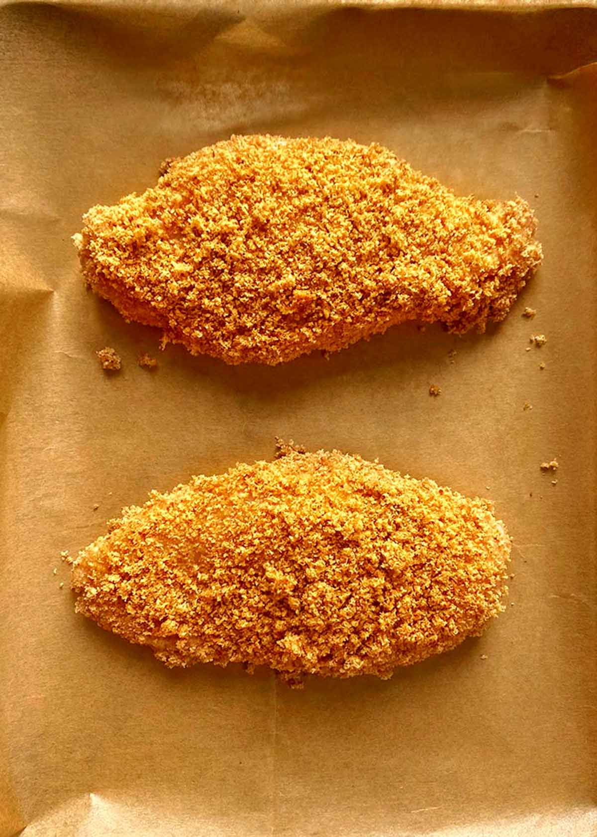 Two breaded chicken breasts on a lined baking tray.