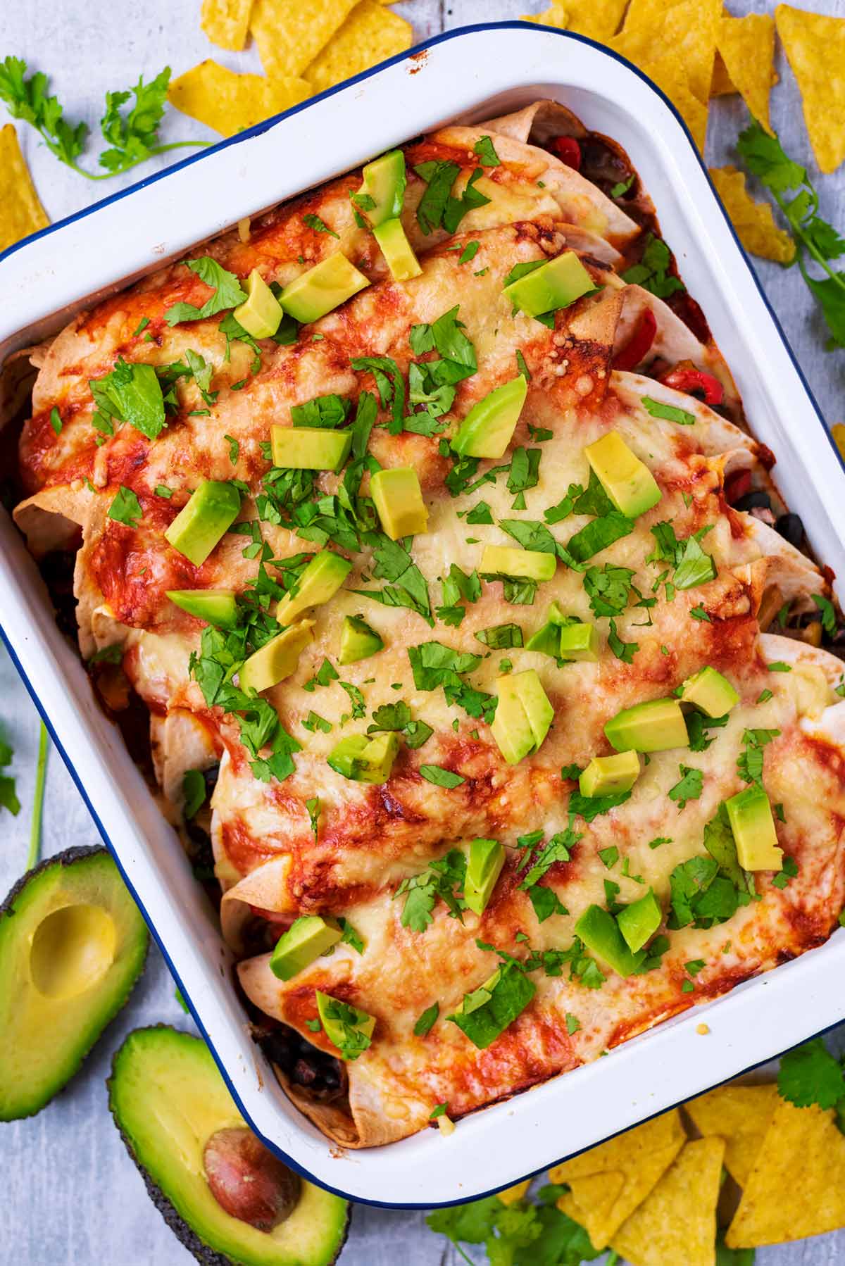 A baking dish full of enchiladas next to tortilla chips and avocado halves.