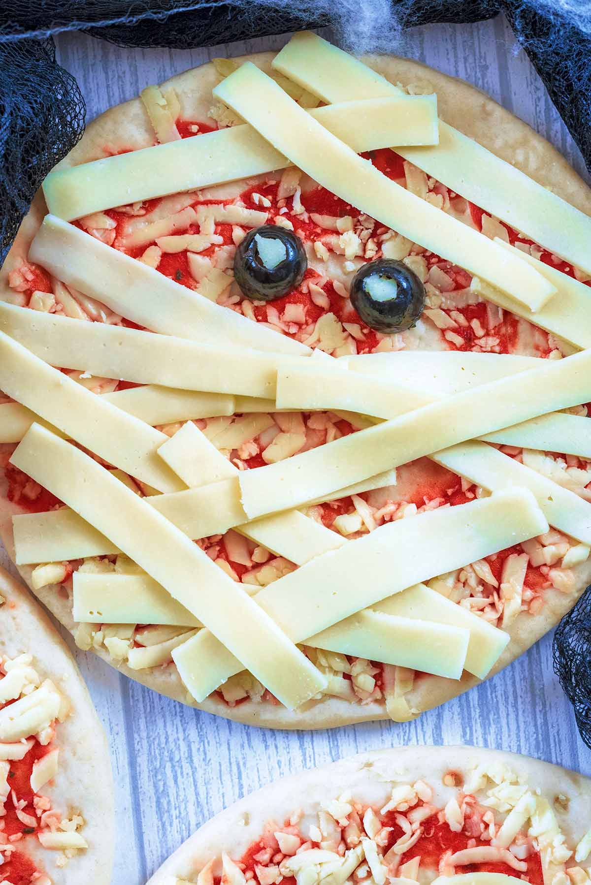 A pizza with strips of cheese across it and two olive halves, making it look like a mummy.