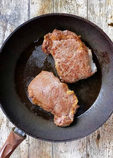 Two large pieces of beef searing in a frying pan.