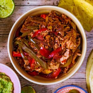 A bowl of slow cooker chicken fajita filling next to guacamole and tortilla wraps.