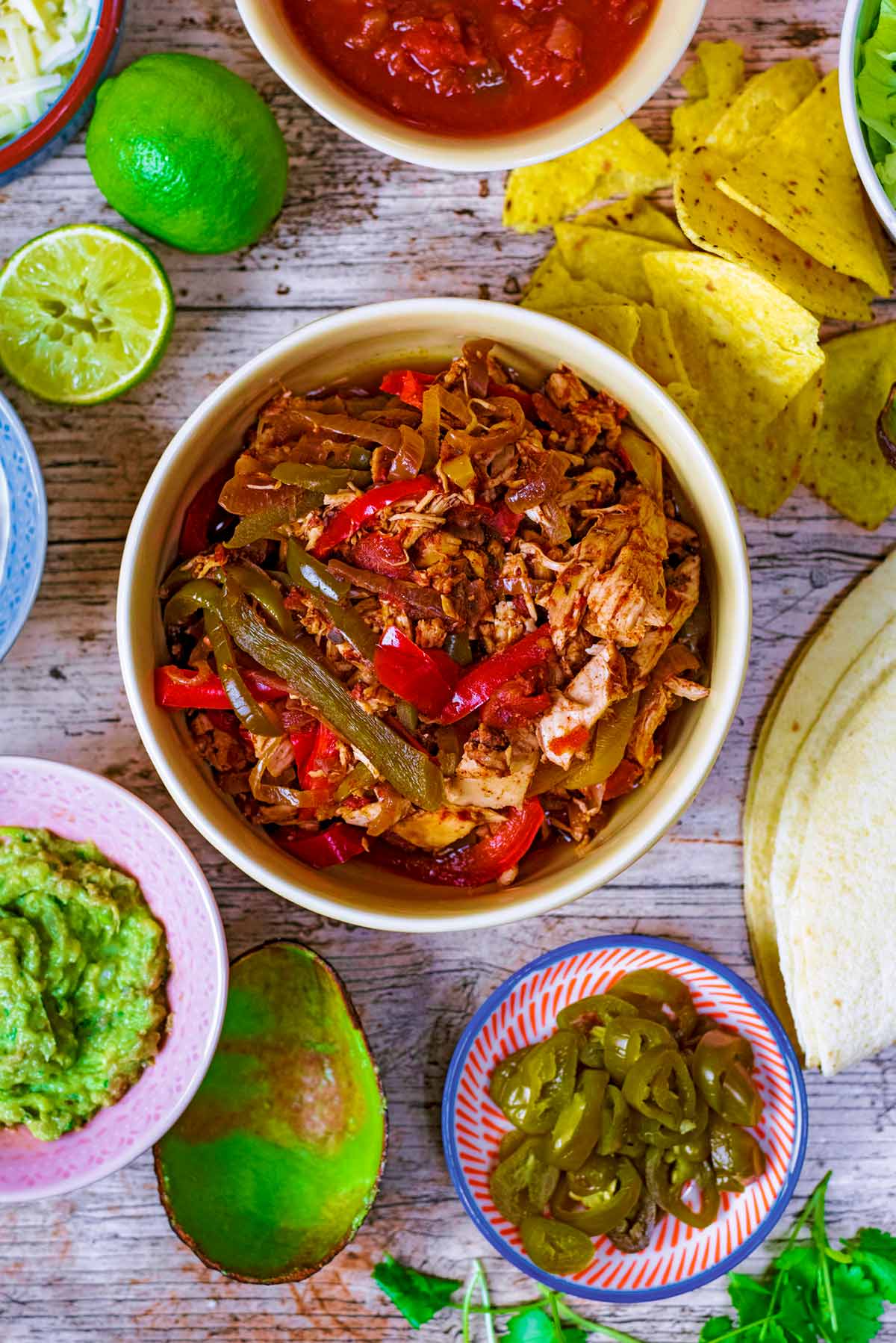A bowl of fajita filling surrounded by salsa, guacamole, tortilla wraps, jalapenos and tortilla chips.