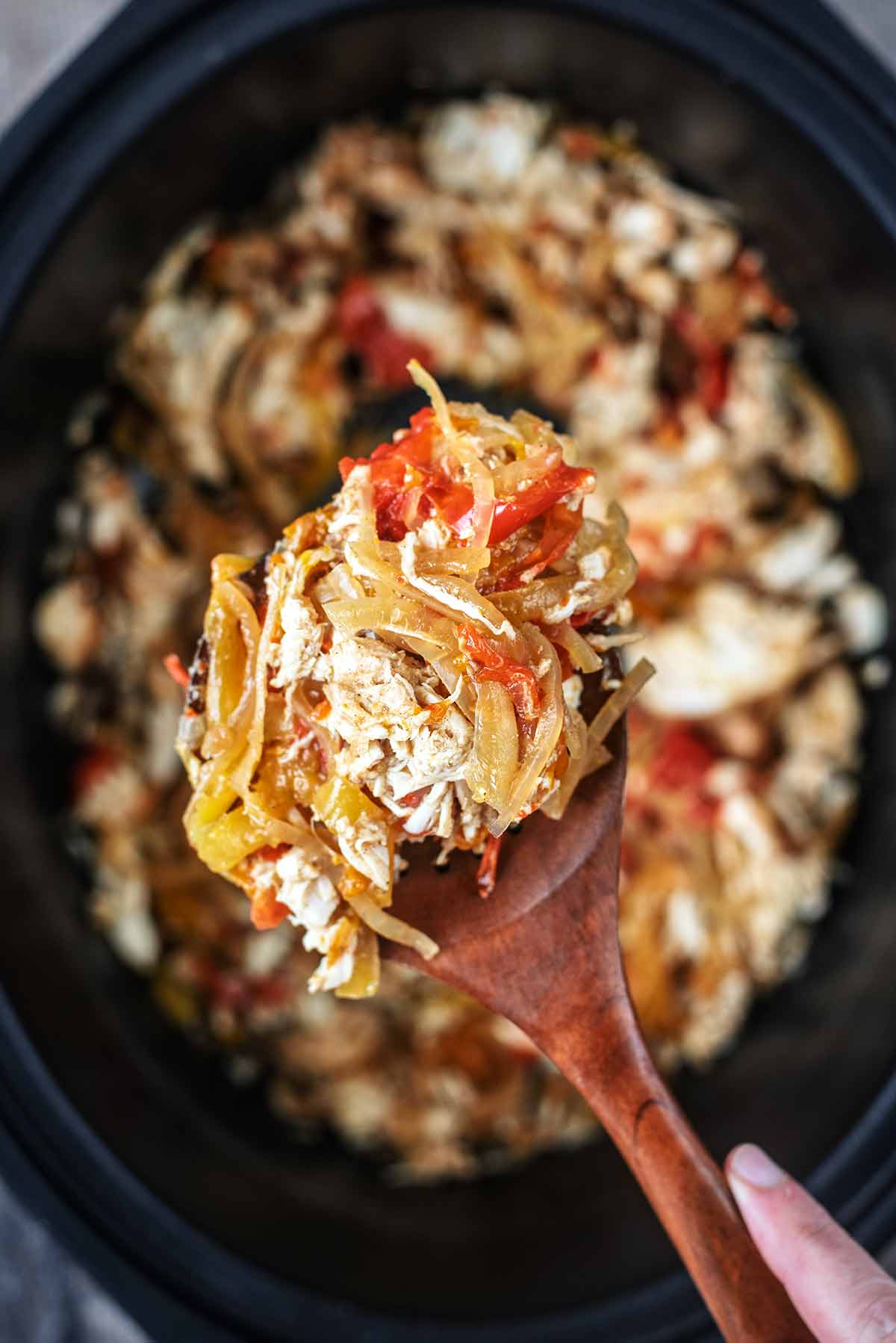 A wooden spoon lifting fajita mixture out of a slow cooker bowl.