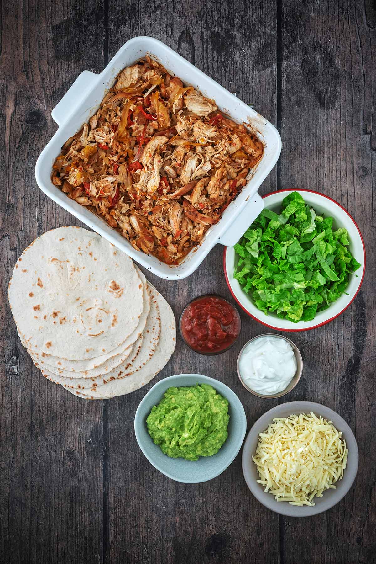 A serving bowl full of fajita mixture next to lettuce, tortilla wraps, salsa, cheese and guacamole.