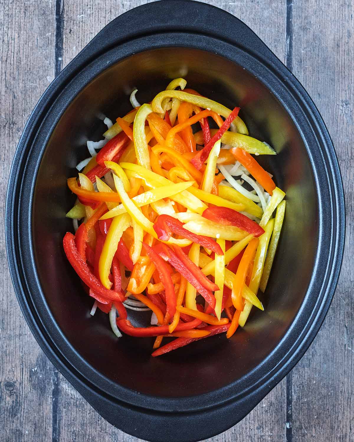Sliced bell peppers and sliced onion in a slow cooker bowl.