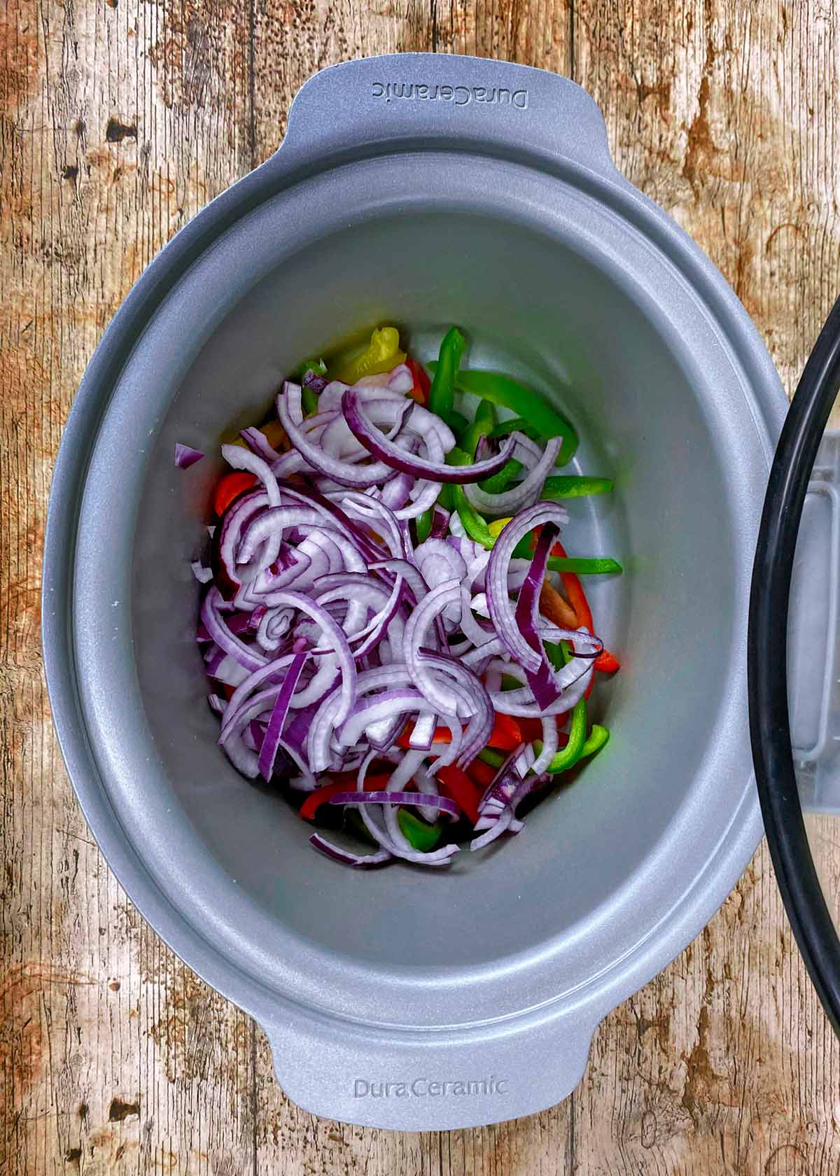 A slow cooker with sliced onions and bell peppers in it.