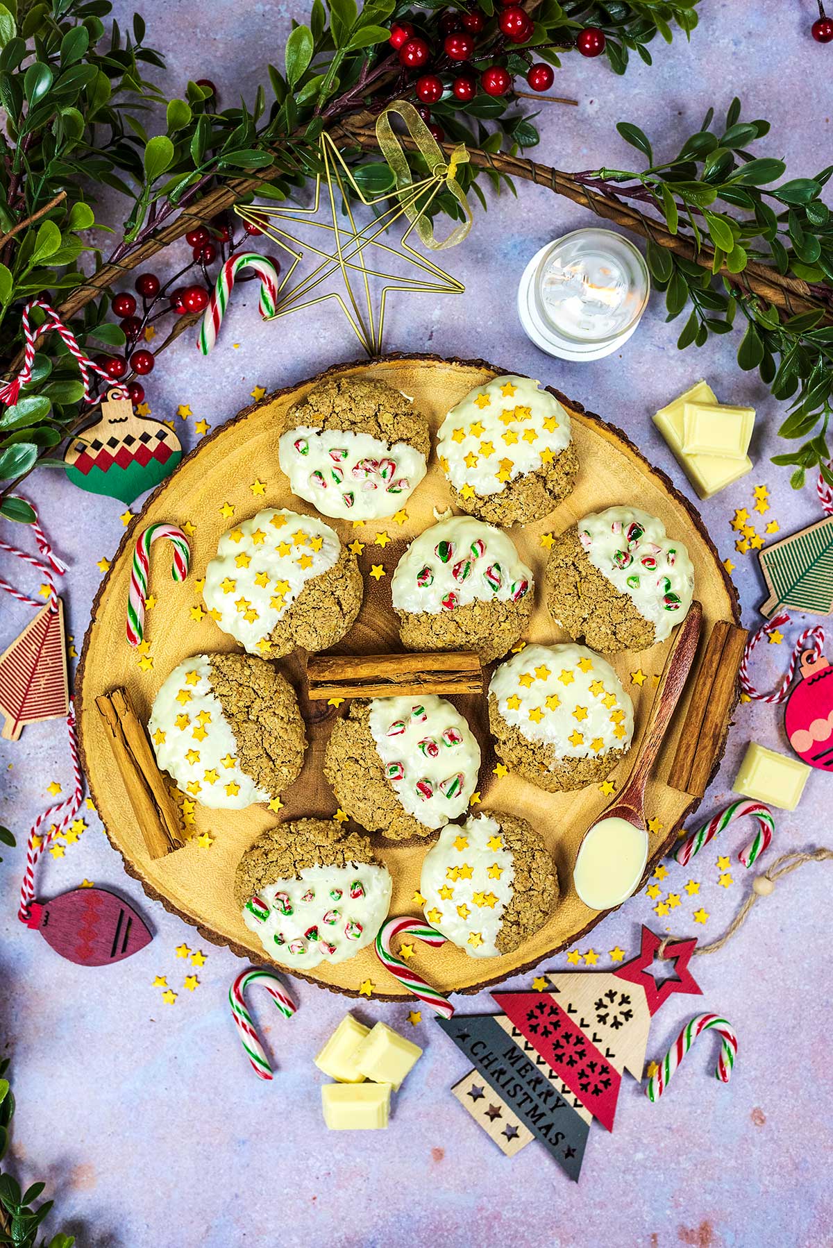 Ten decorated cookies on a round wooden board surrounded by Christmas decorations.