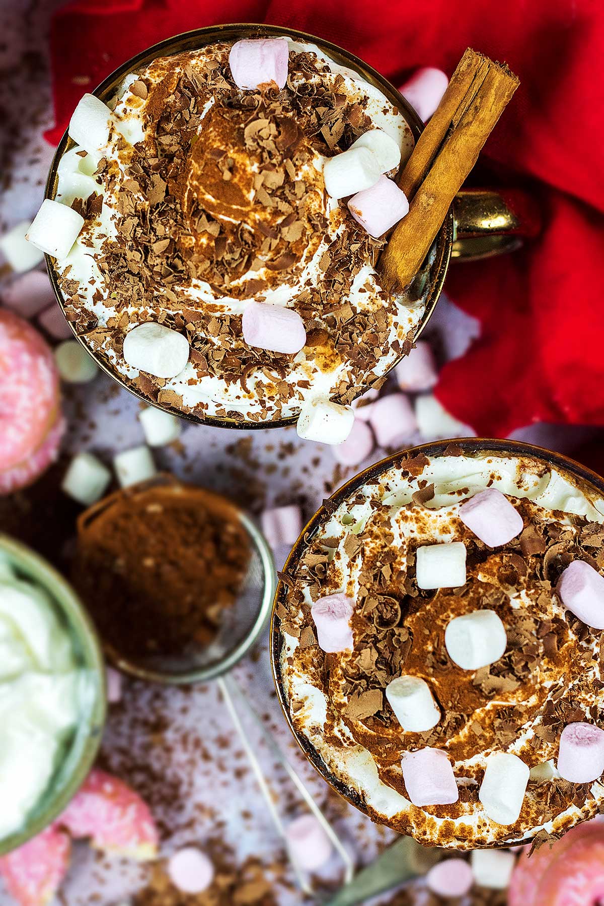 Whipped cream, marshmallows and chocolate shavings on top of two mugs of hot chocolate as viewed from above.