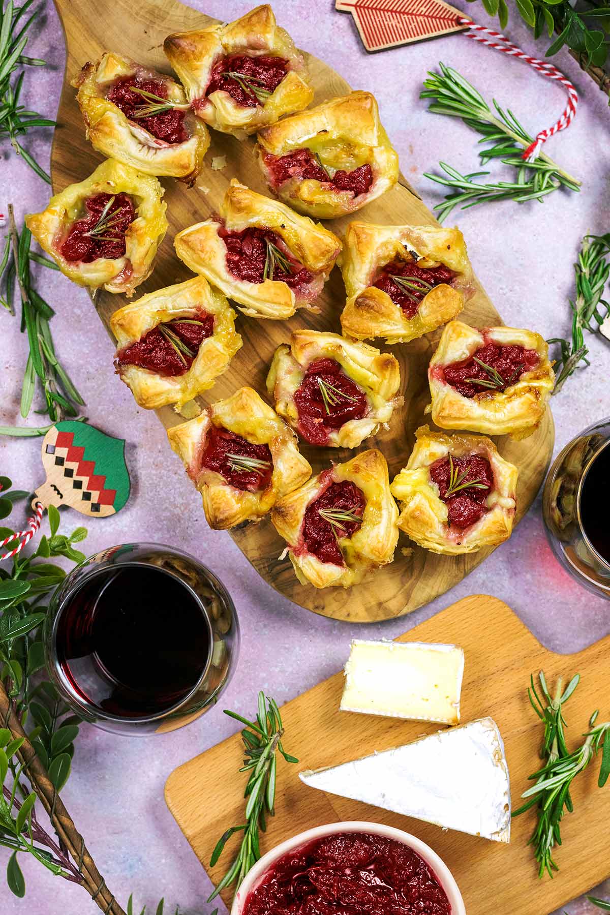 Brie and cranberry bites on a serving board next to some glasses of red wine and a board with cheese on it.