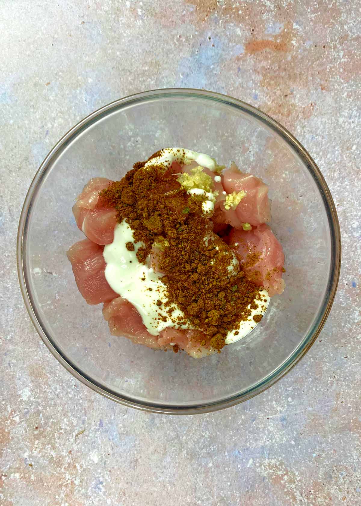 A glass bowl containing chunks of chicken, yogurt and a spice blend.