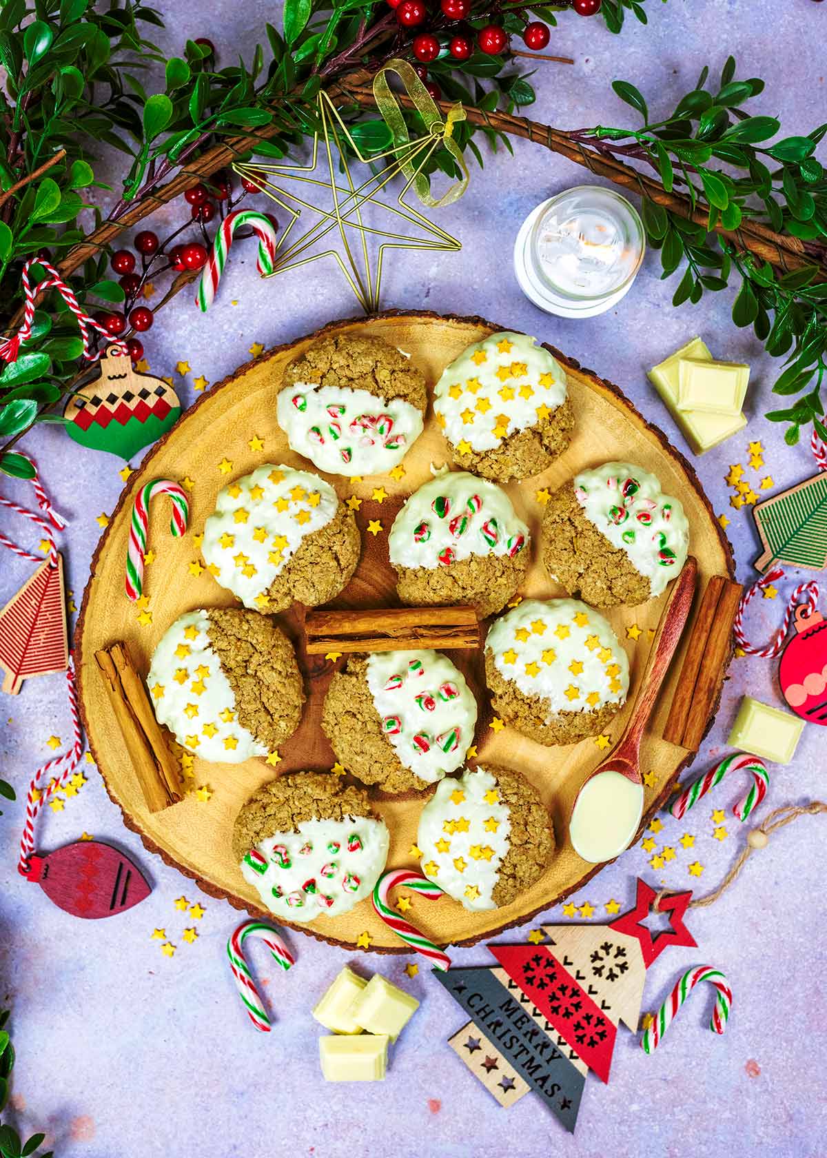 Ten decorated cookies on a round wooden board surrounded by Christmas decorations.
