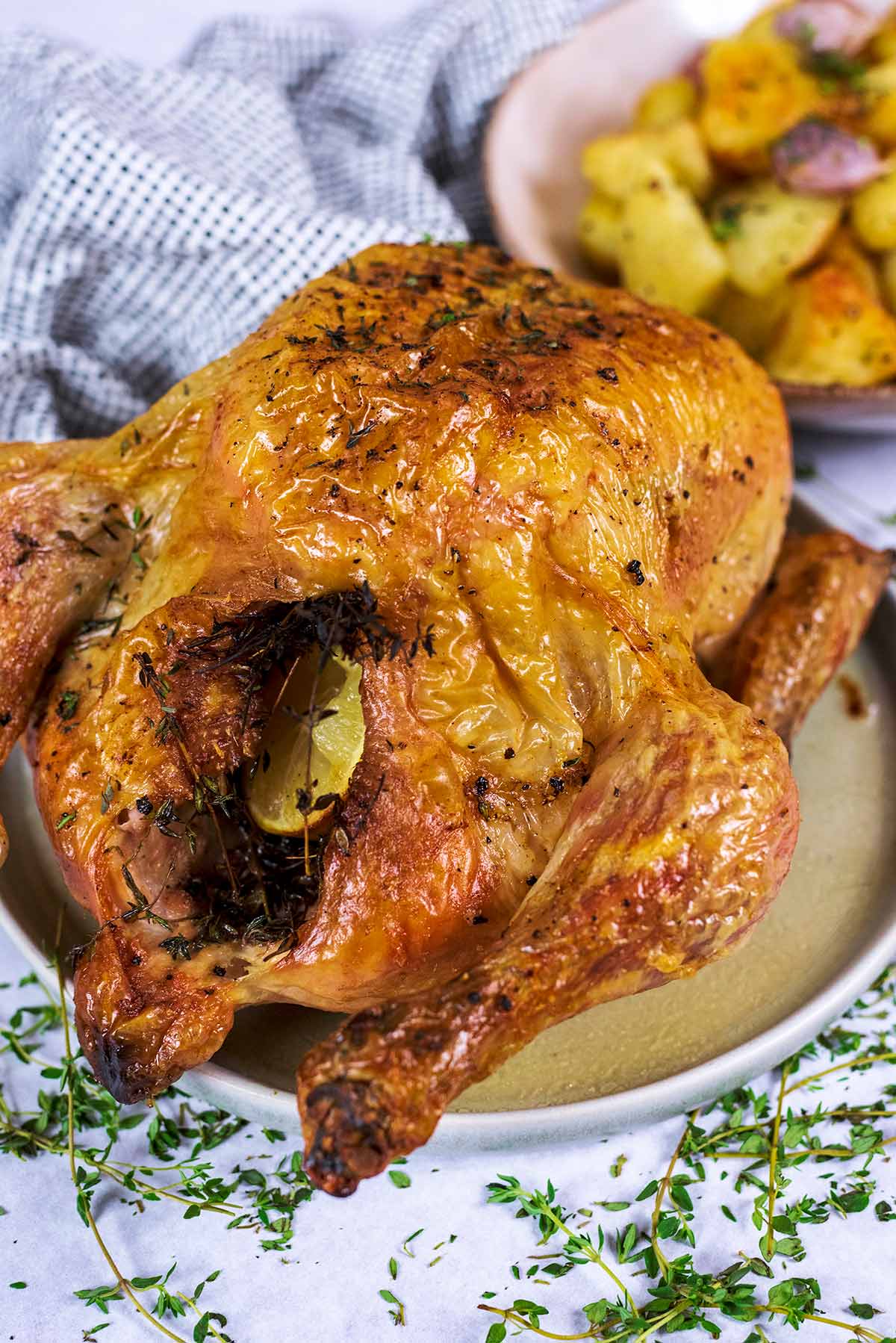 A whole roast chicken on a plate in front of a bowl of roasted potatoes.