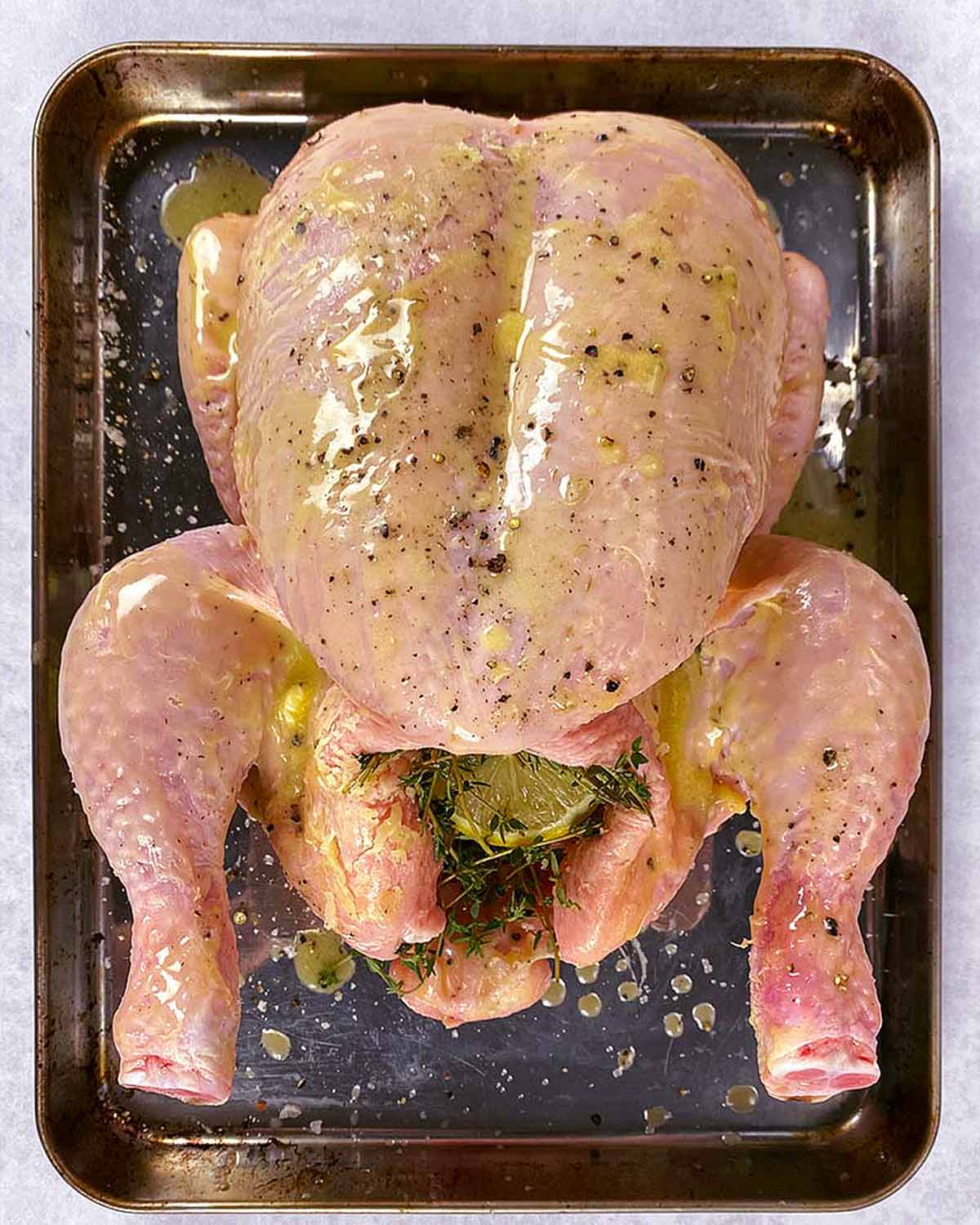A whole uncooked chicken on a baking tray with lemons and thyme in its cavity.