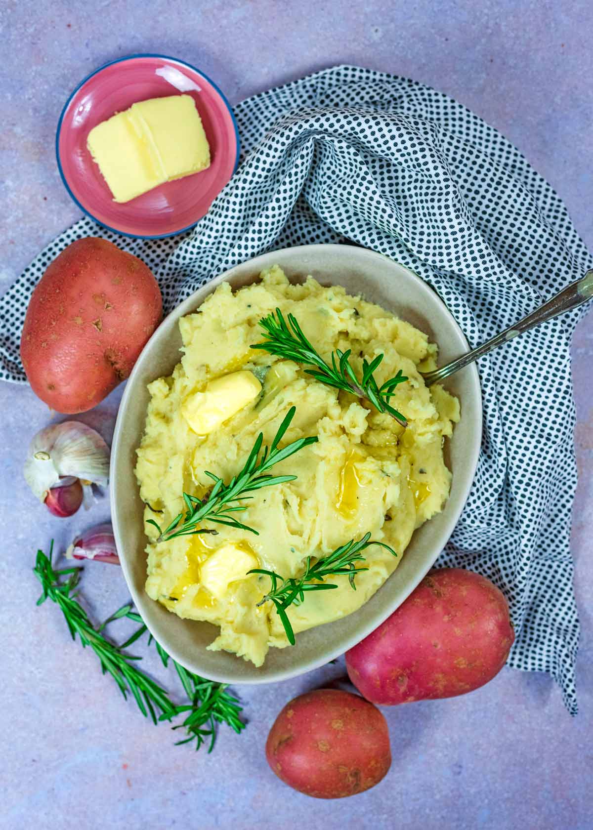 A bowl of mashed potato surrounded by whole potatoes, butter, garlic and rosemary.