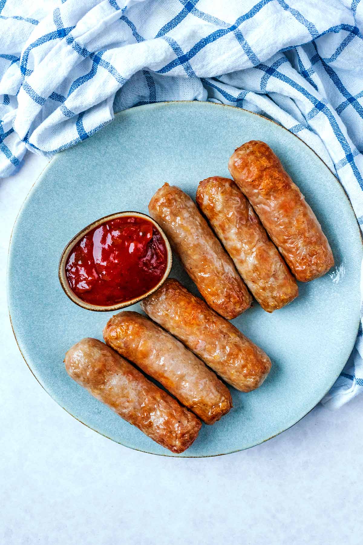 Six cooked sausages on a round plate with a small pot of ketchup.