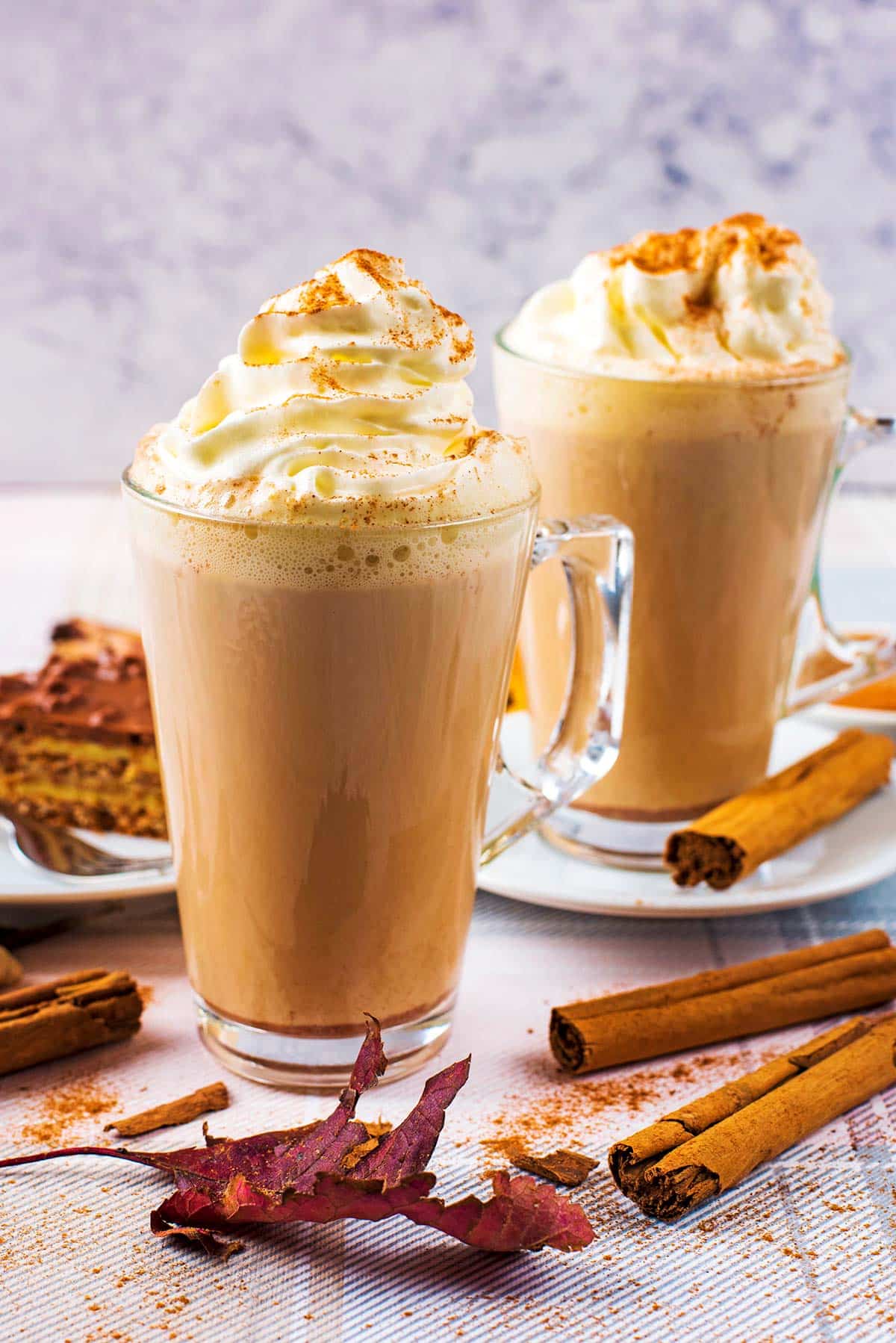 Two glasses of milky coffee topped with whipped cream. Cinnamon sticks lay next to them.