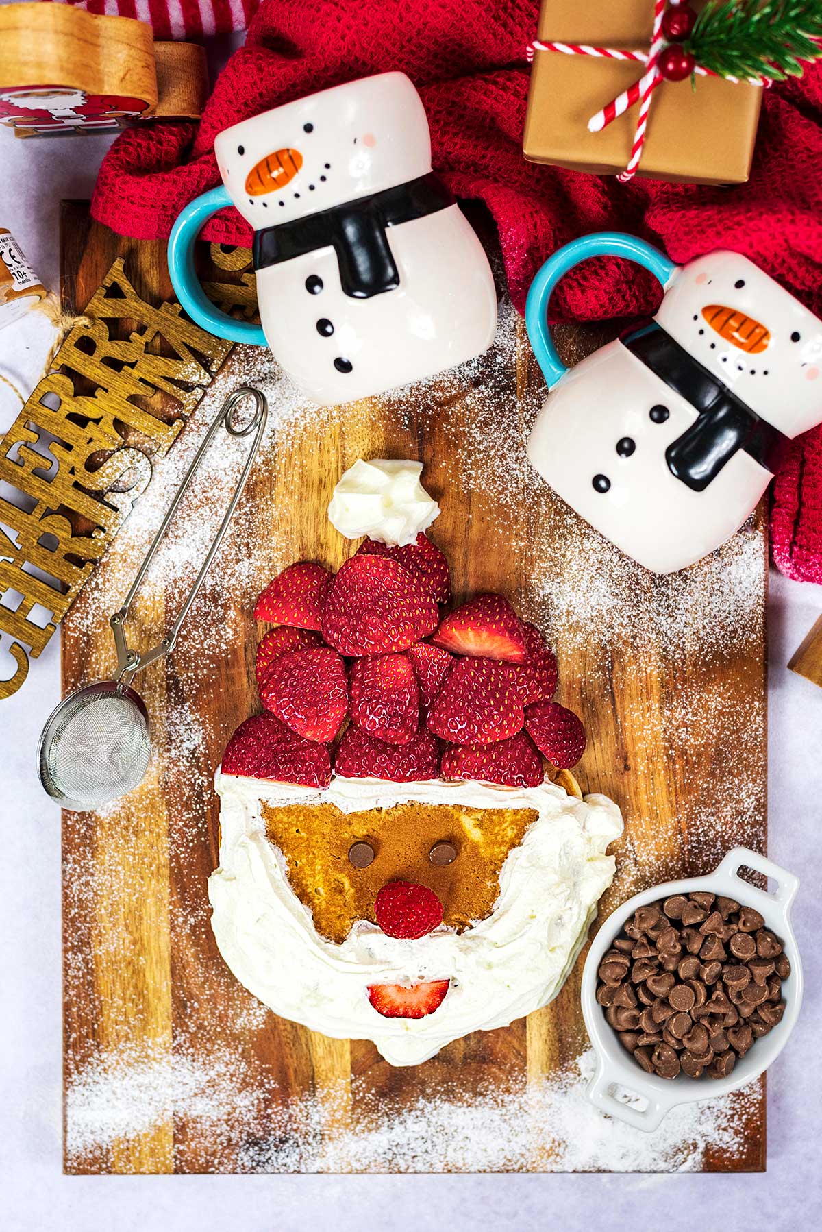A pancake decorated like Santa Claus next to two snowman shaped mugs.