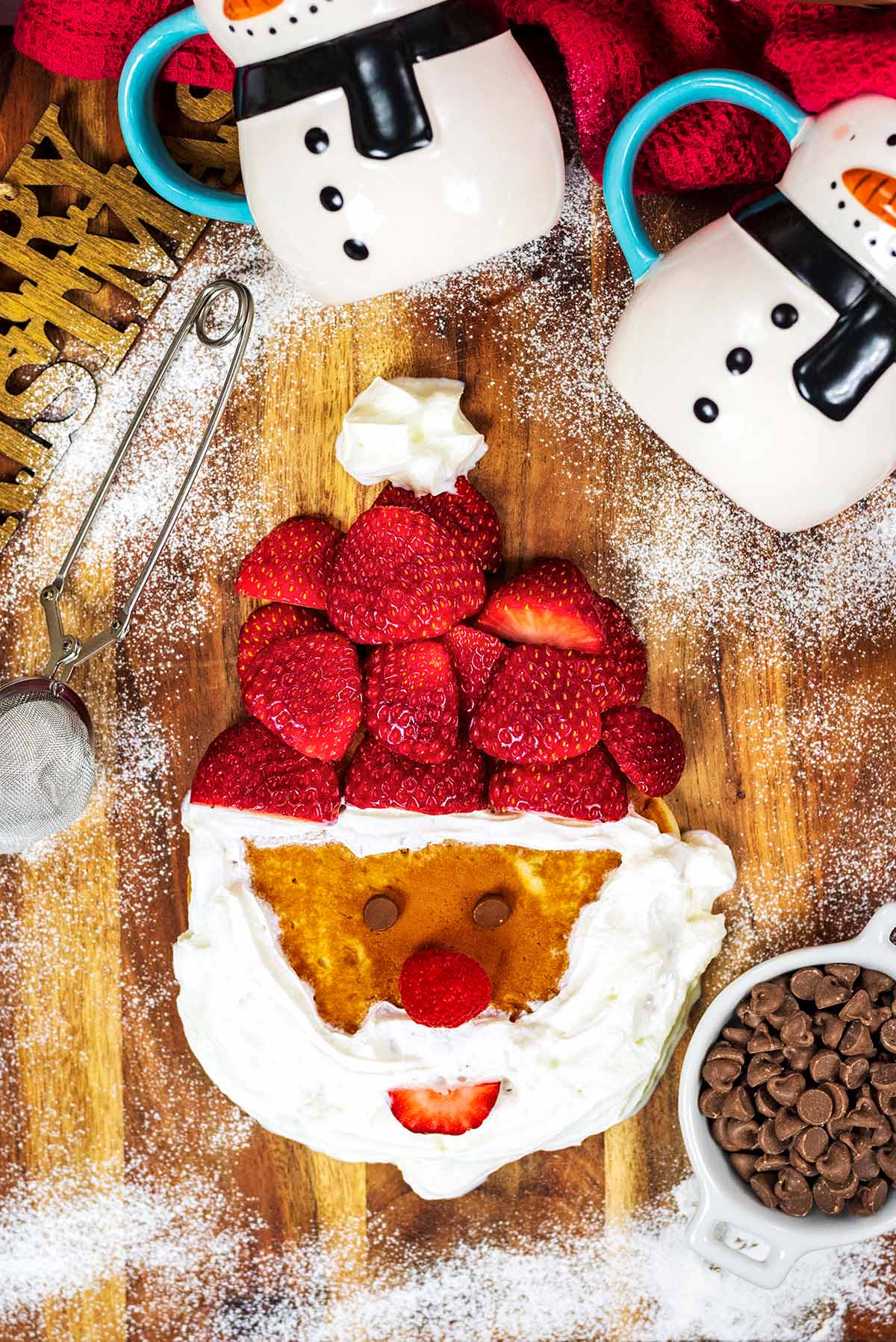 A pancake decorated like Santa Claus next to two snowman shaped mugs.